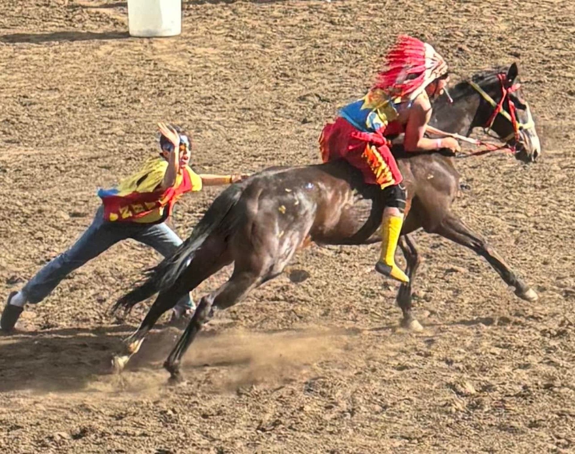 Horse race on dirt track with rider in feathered headdress and running handler.