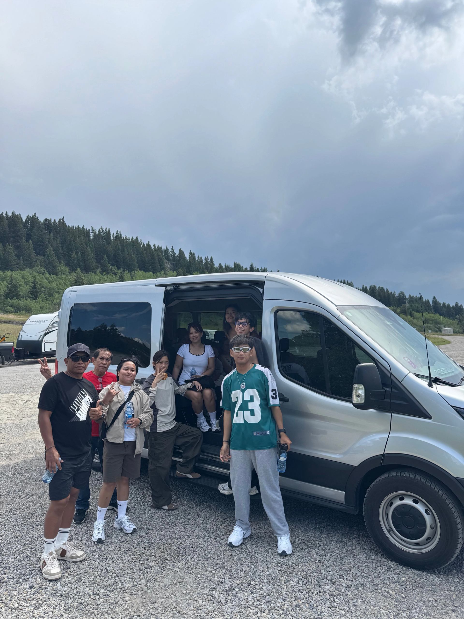 Group of people standing by a parked silver van on gravel near forested hills.