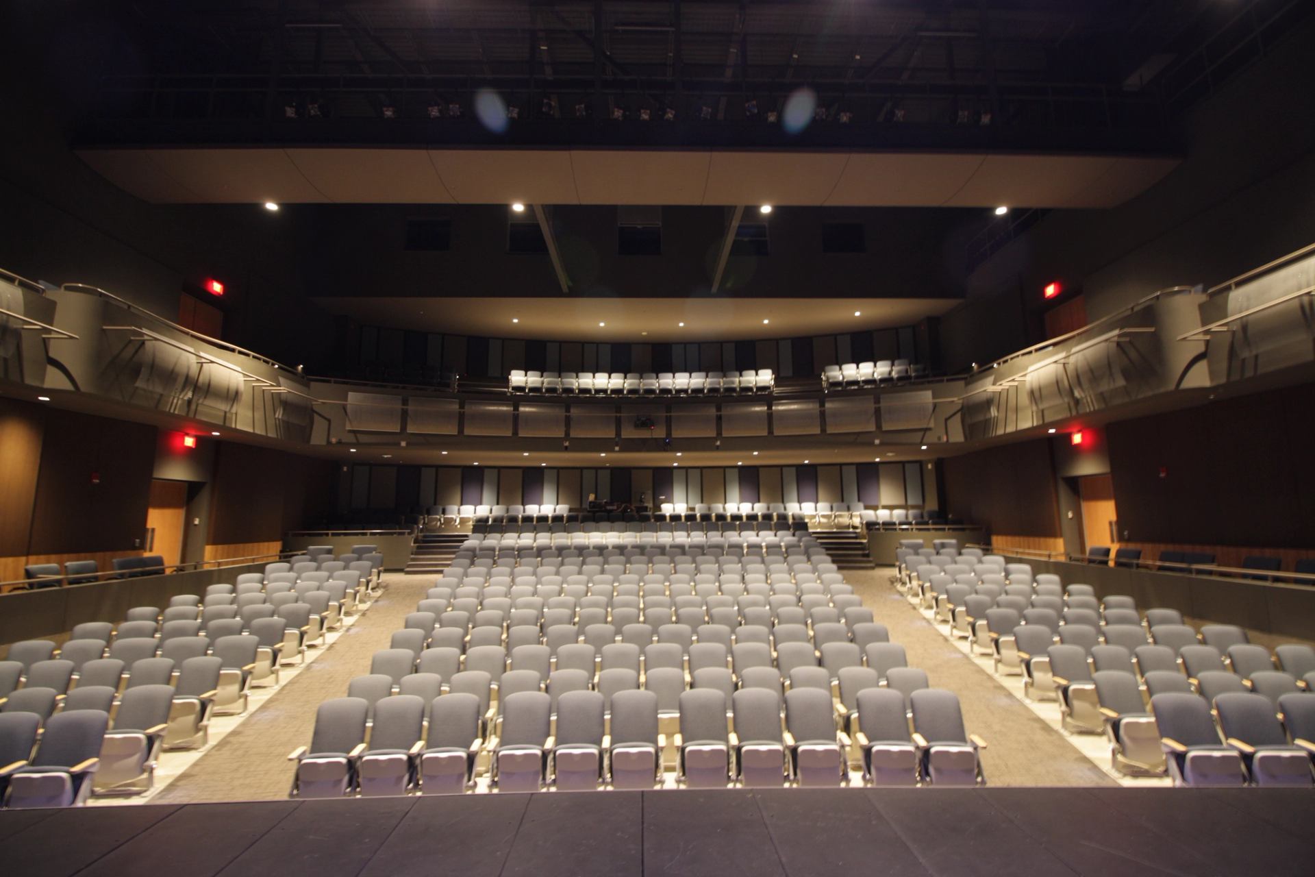 Empty theatre auditorium with rows of grey seats and upper balcony under stage lights.