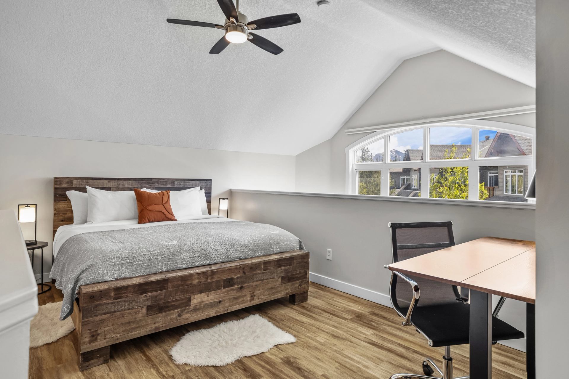 Bedroom with wood bed, desk, office chair, vaulted ceiling and large arched window.