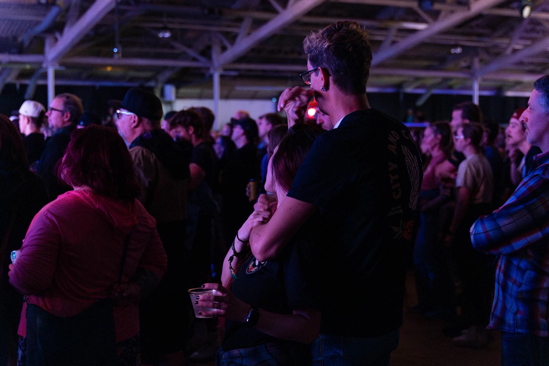 Crowd stands indoors at a concert, watching the stage under purple lighting.