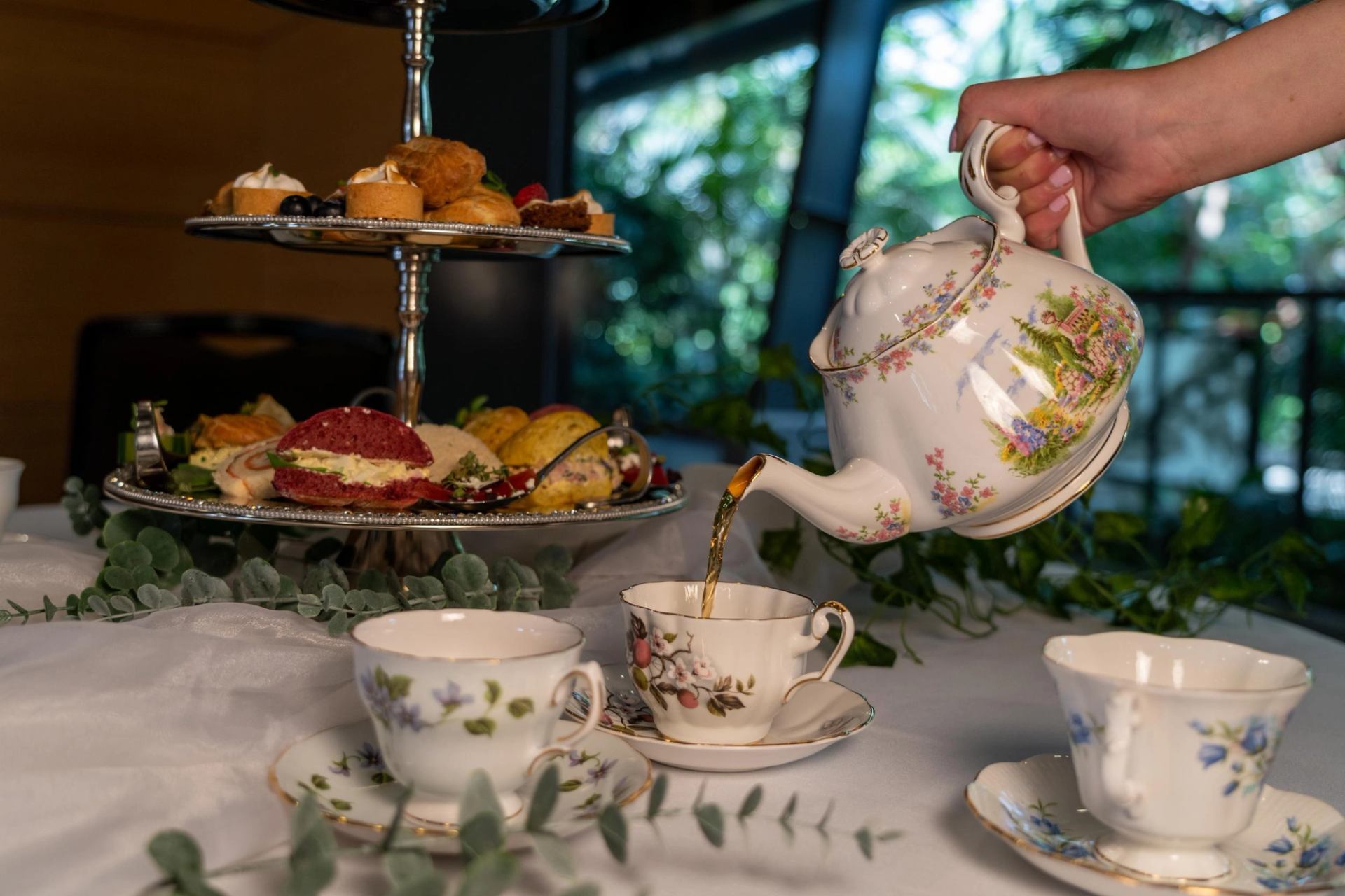 A hand pours tea from a floral teapot into a teacup, next to a tiered stand of pastries and sandwiches.