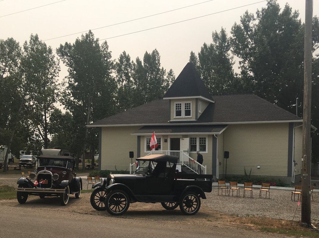 Two vintage vehicles are parked outside a heritage building surrounded by trees and seating.