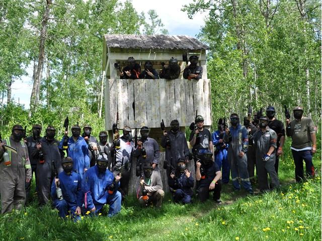 Paintball players in camouflage gear and masks pose in front of a wooden shooting station.