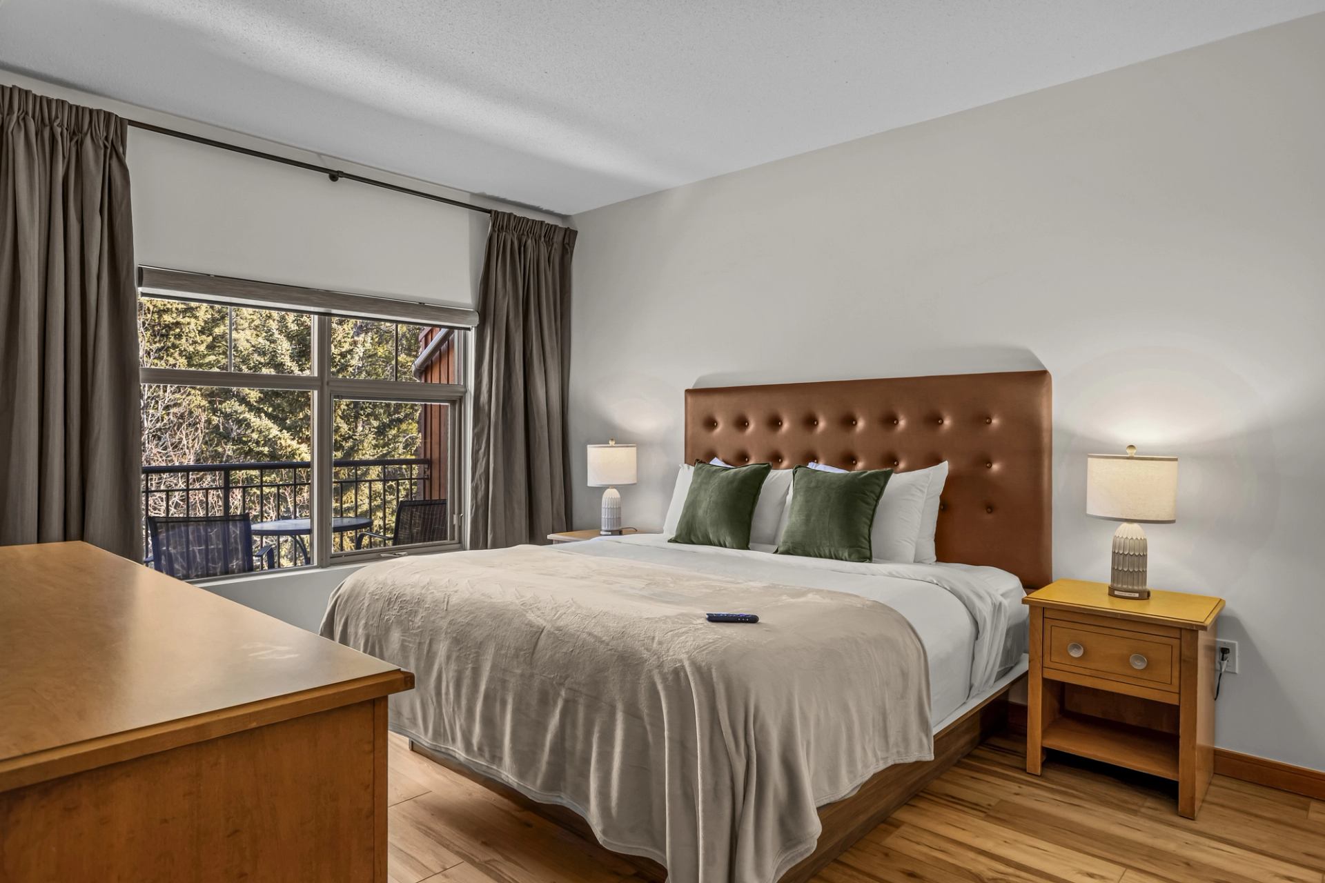 A spacious bedroom includes a large bed, bedside tables with lamps, and a window leading to a balcony with forest views.