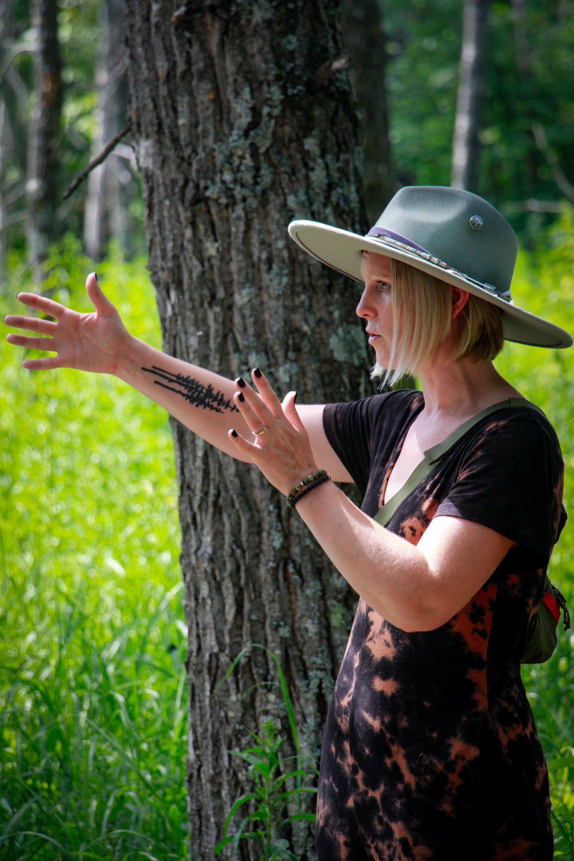 A blonde woman in a green hat and tie-dye shirt gestures with an arm featuring a forest silhouette tattoo, outdoors by a tree.