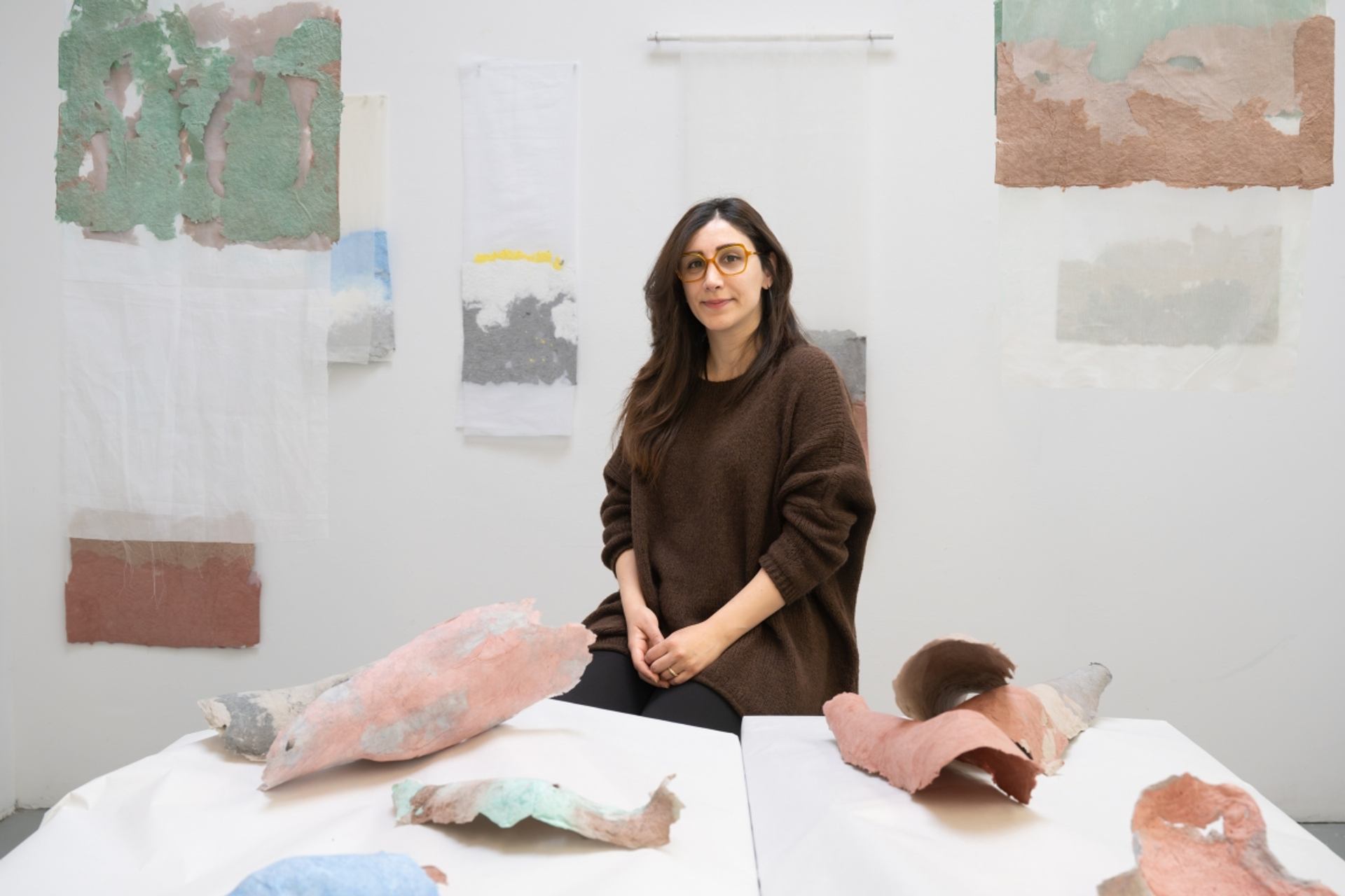 Artist sits in studio surrounded by abstract textured paper sculptures and wall hangings.