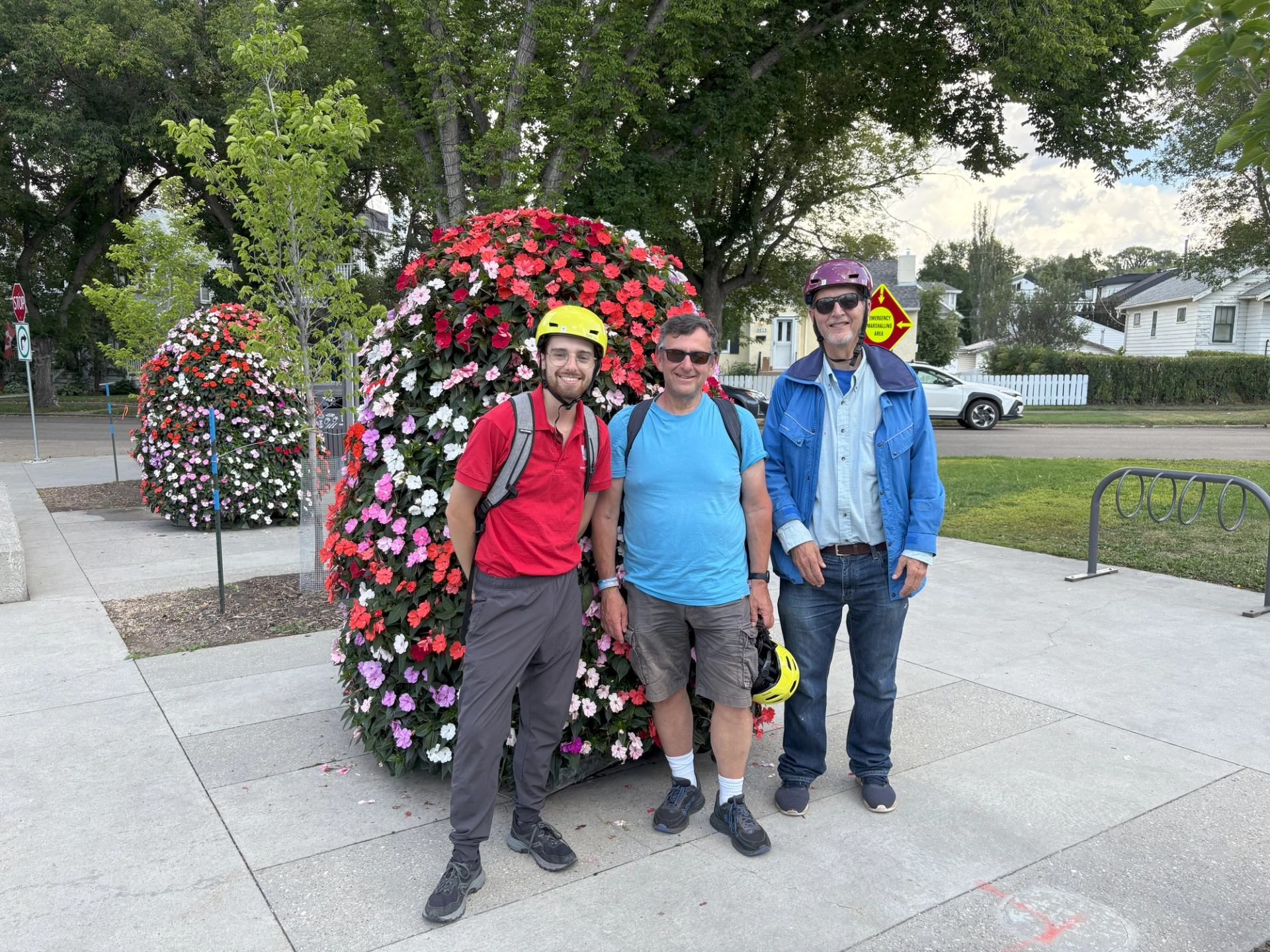 Three people stand by a large floral sphere, two wearing helmets, with trees and buildings behind.