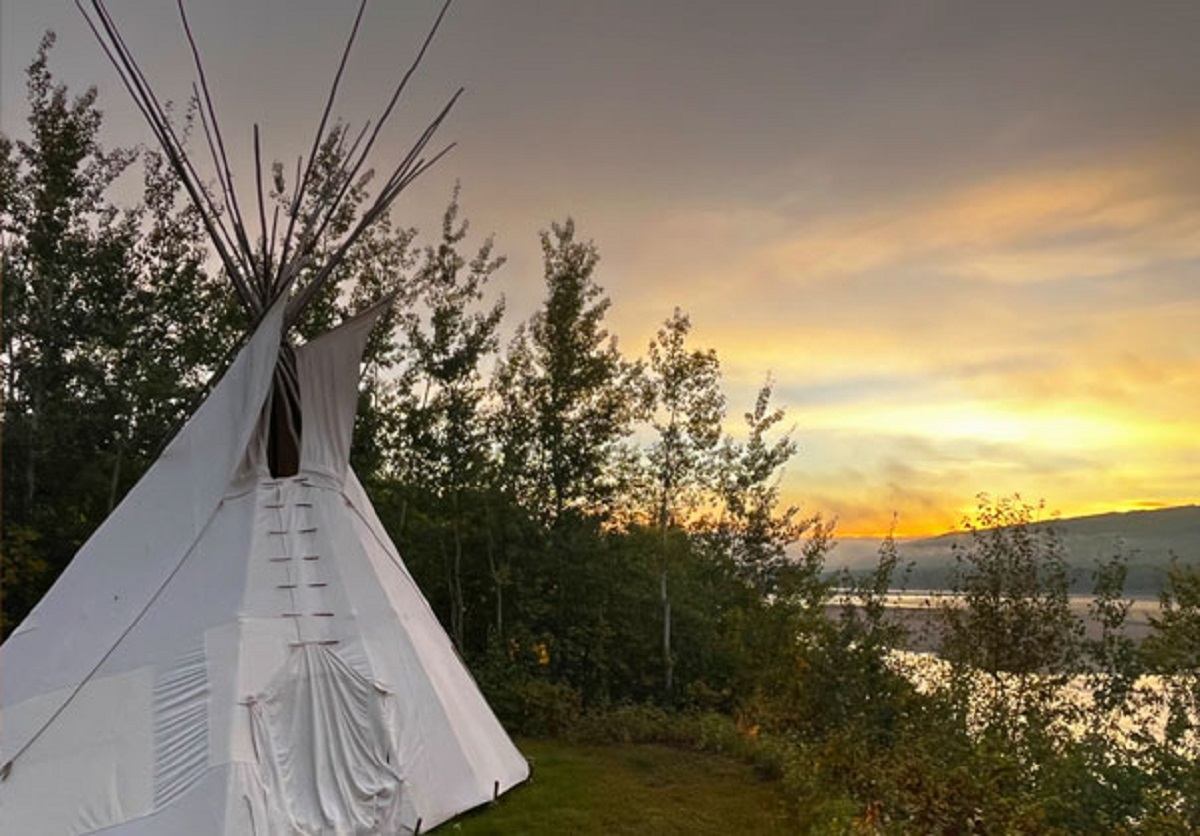 A tranquil tipi by the river, surrounded by trees and bathed in sunset light.