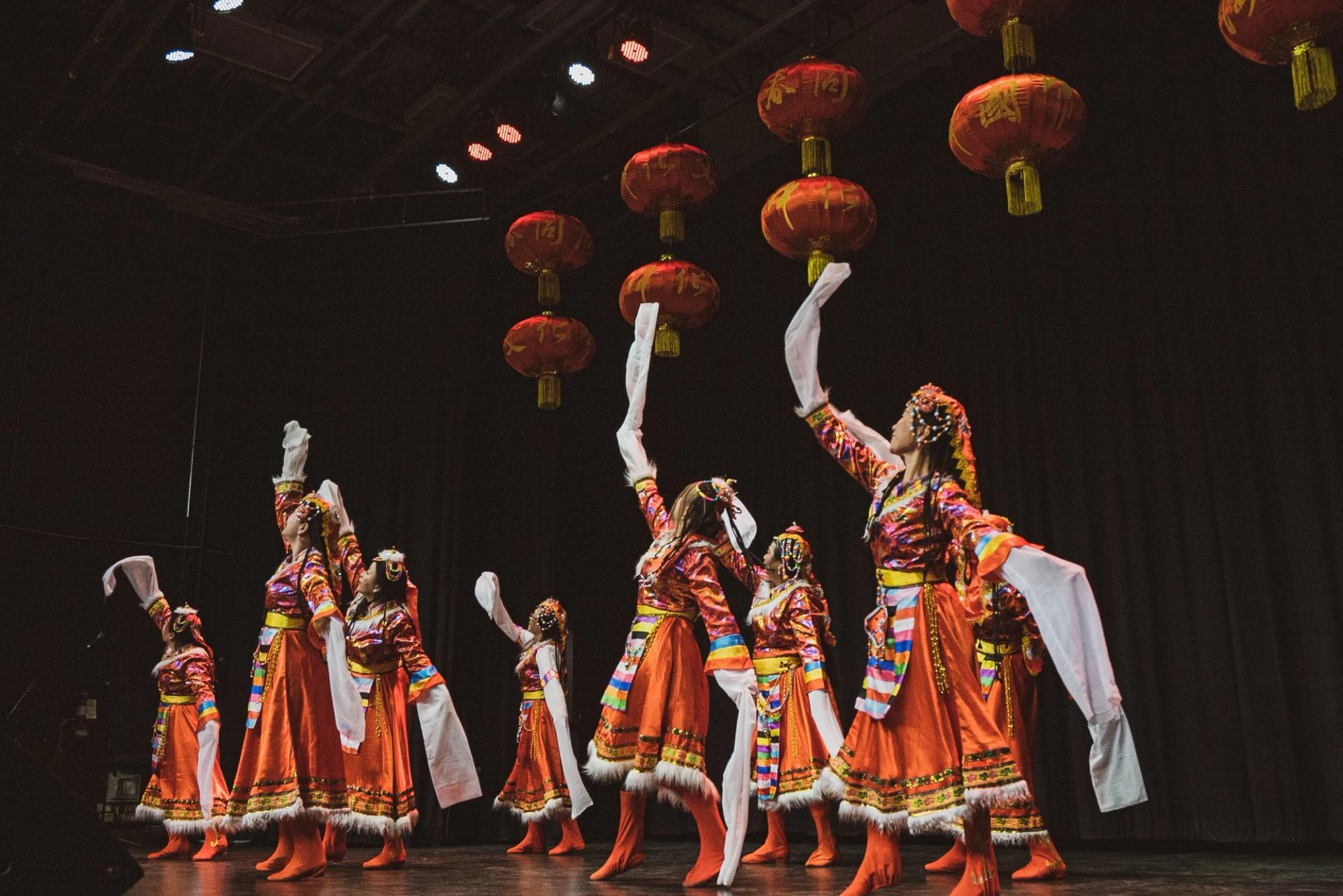 Traditional dancers in vibrant orange costumes performing under hanging red lanterns on stage.