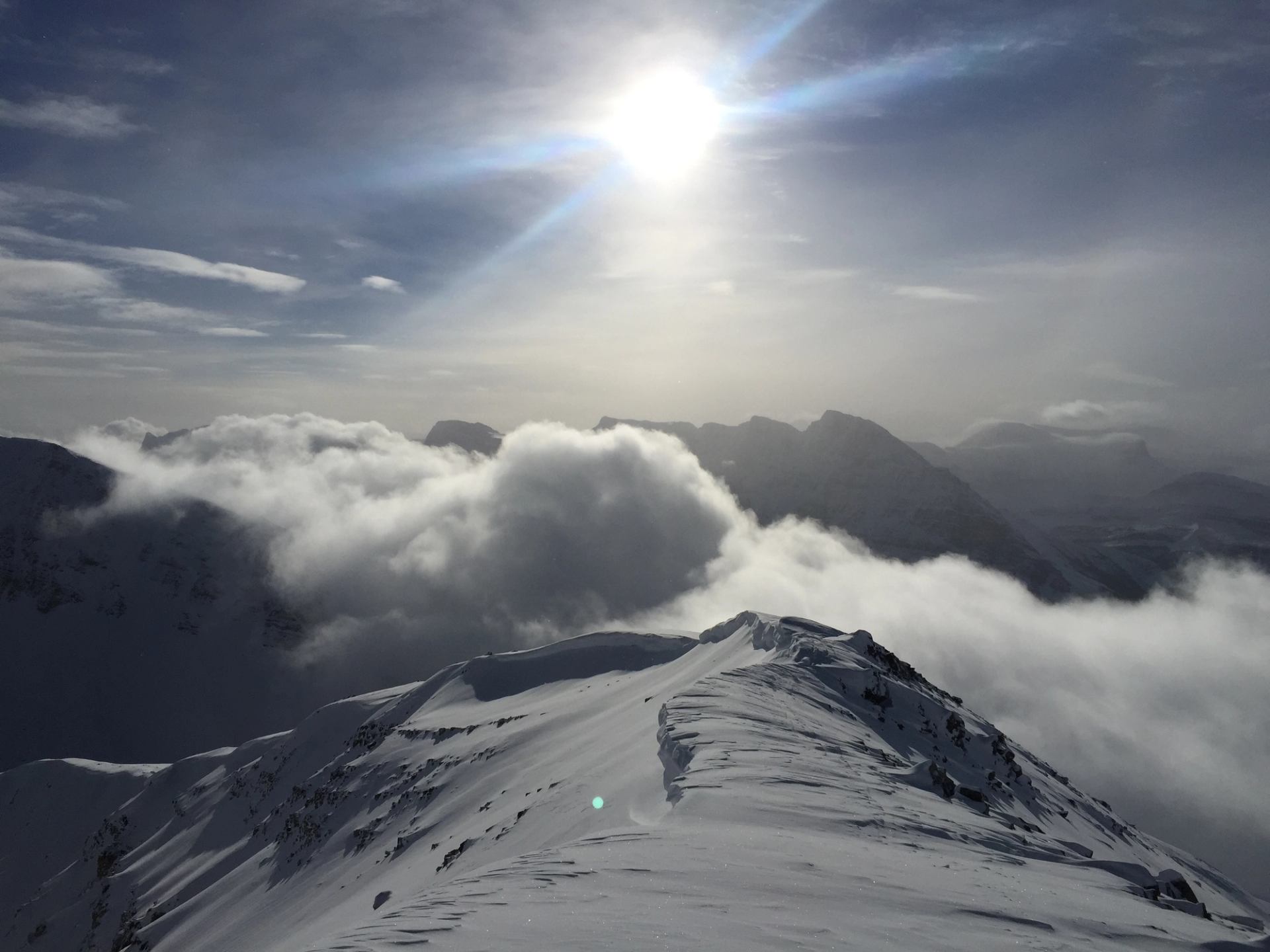 Snow-covered mountain ridge above clouds under bright sun and hazy distant peaks.