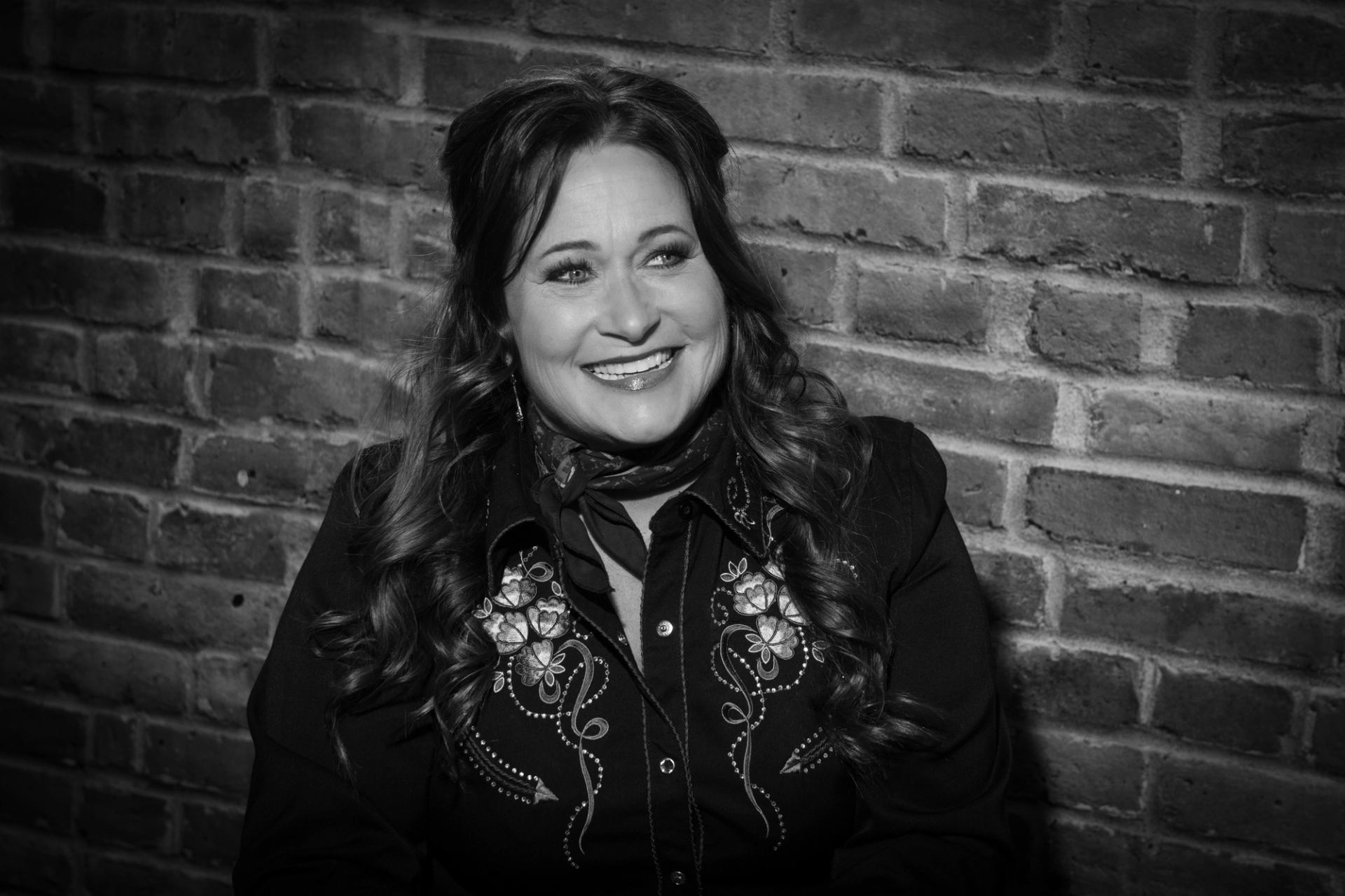 Woman with long wavy hair sits by brick wall, wearing embroidered western shirt.