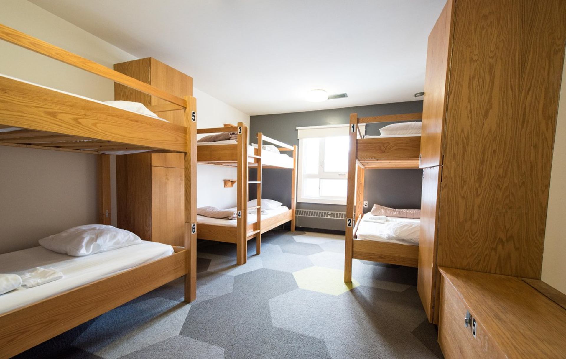 Shared dorm room at HI Calgary with wooden bunk beds, numbered bunks, seating area with green chairs.