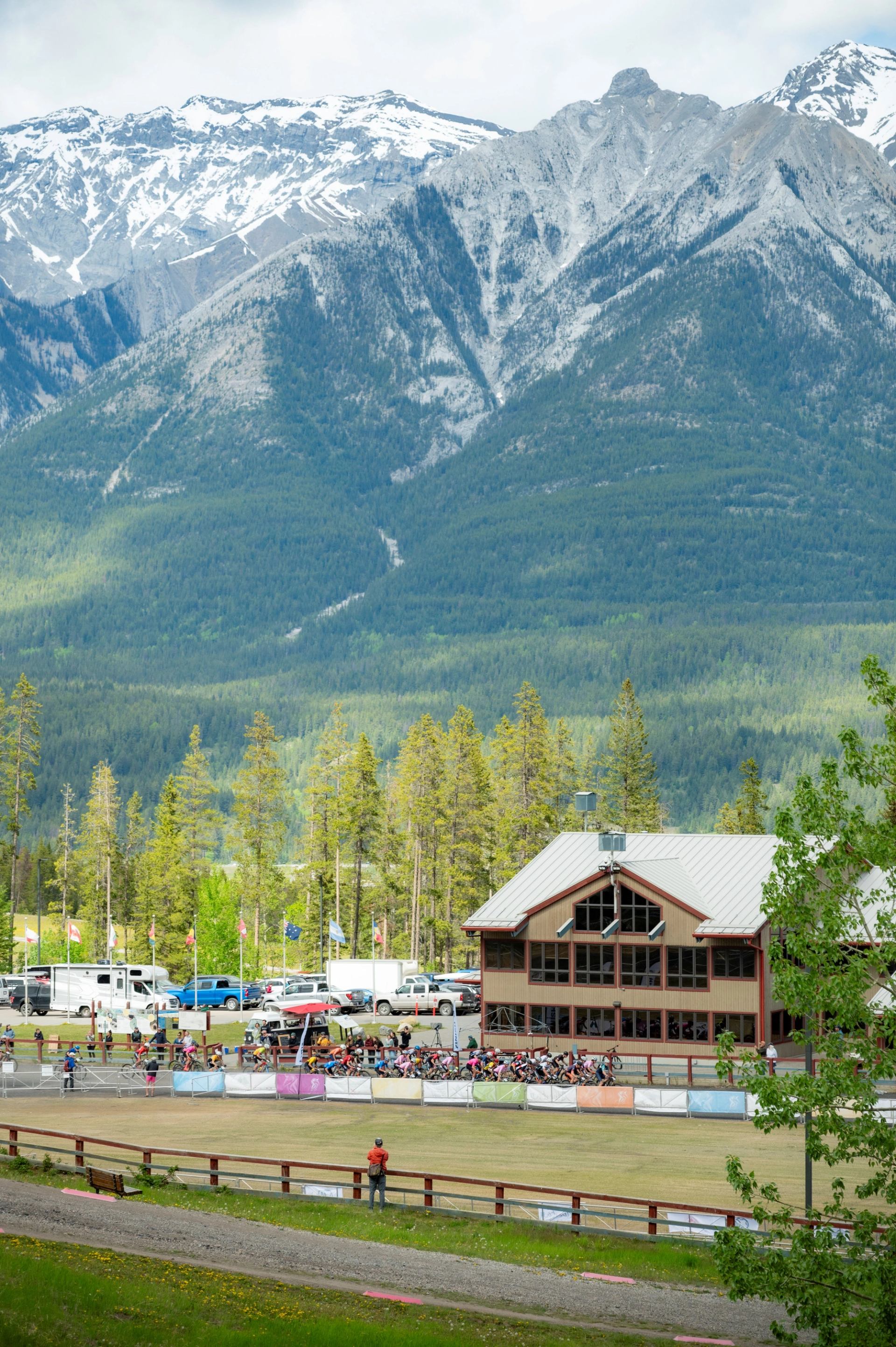 Mountain bike race venue with a lodge, spectators, and fencing framed by forested slopes and rocky mountain peaks.