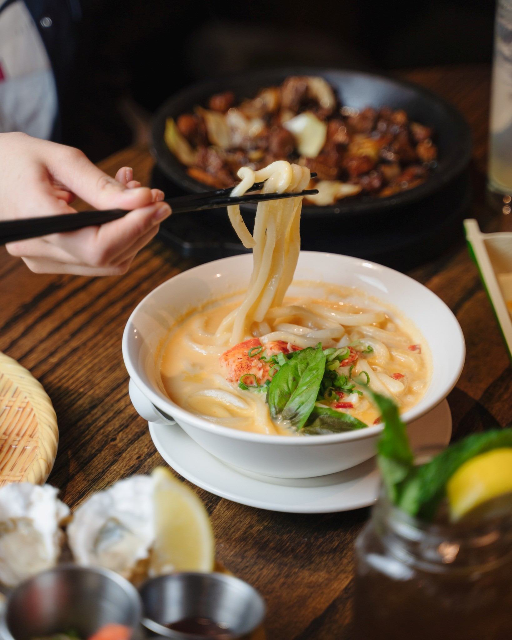 Bowl of noodles with chopsticks lifting strands, garnish and herbs on wooden table.