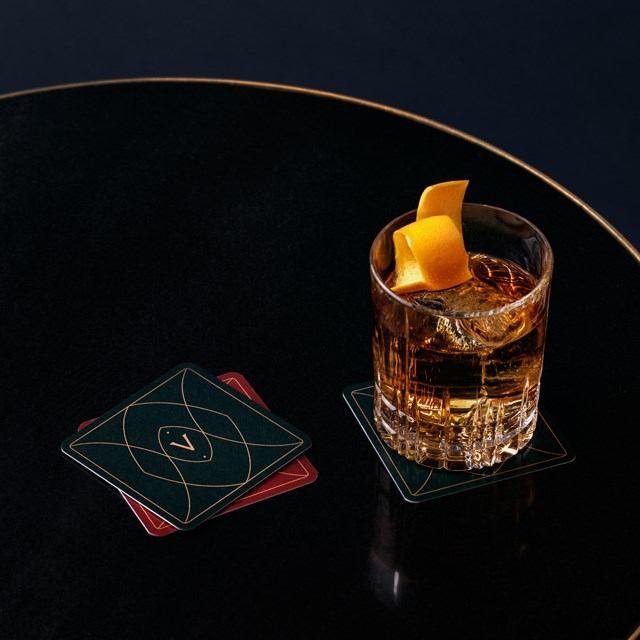 Whiskey cocktail with orange peel garnish on a black table beside decorative coasters.