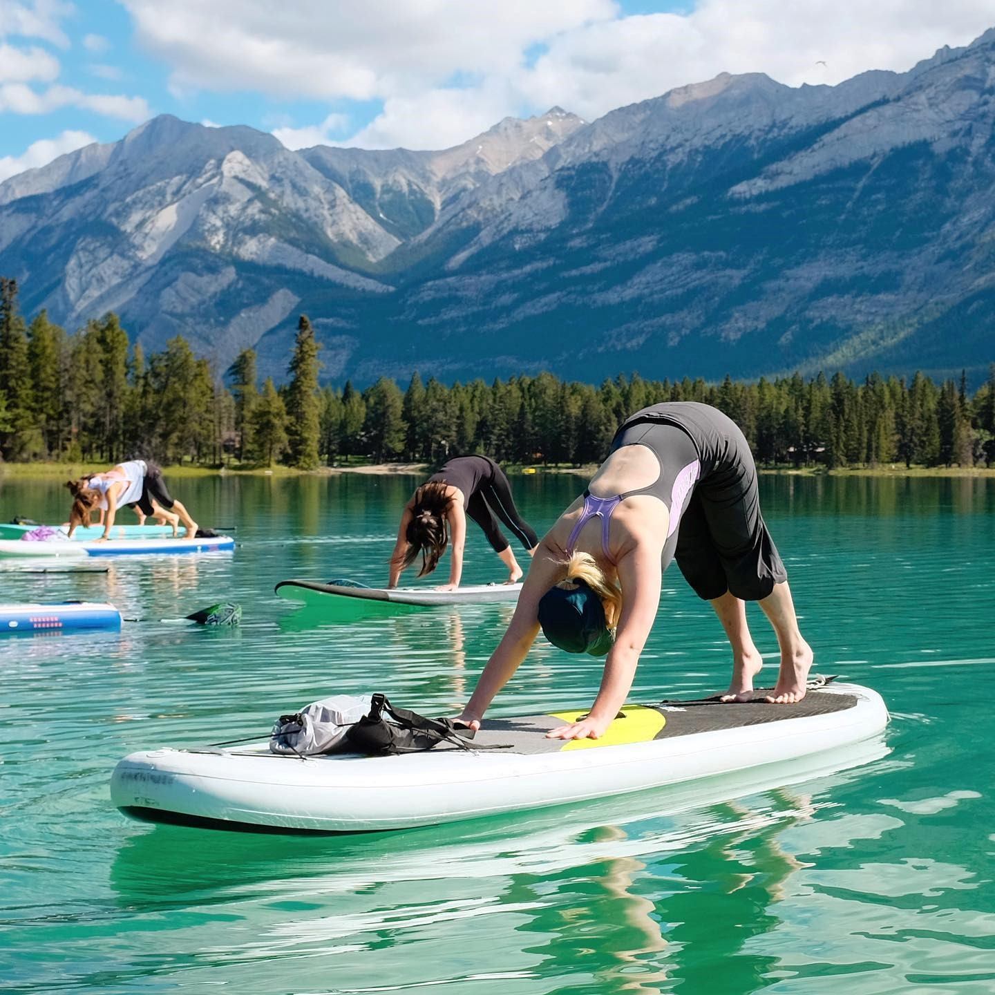 People practise yoga on paddleboards on turquoise lake, mountains and trees behind.