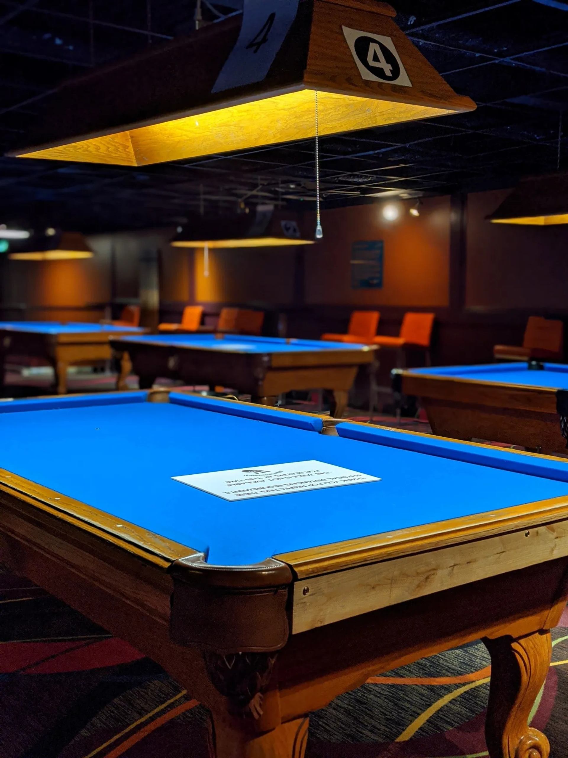 Pool hall with blue felt tables, overhead lights, and orange chairs in background.