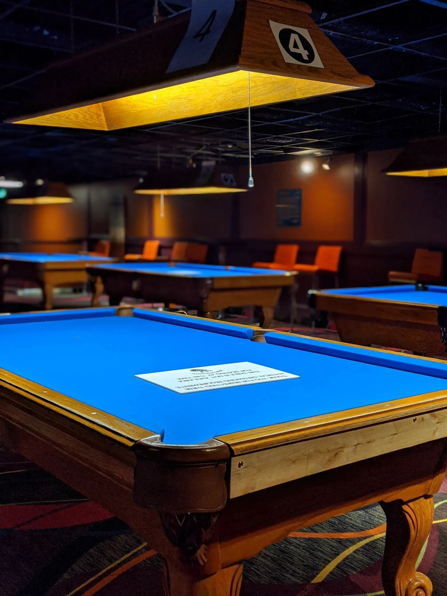 Pool hall with blue felt tables, overhead lights, and orange chairs in background.