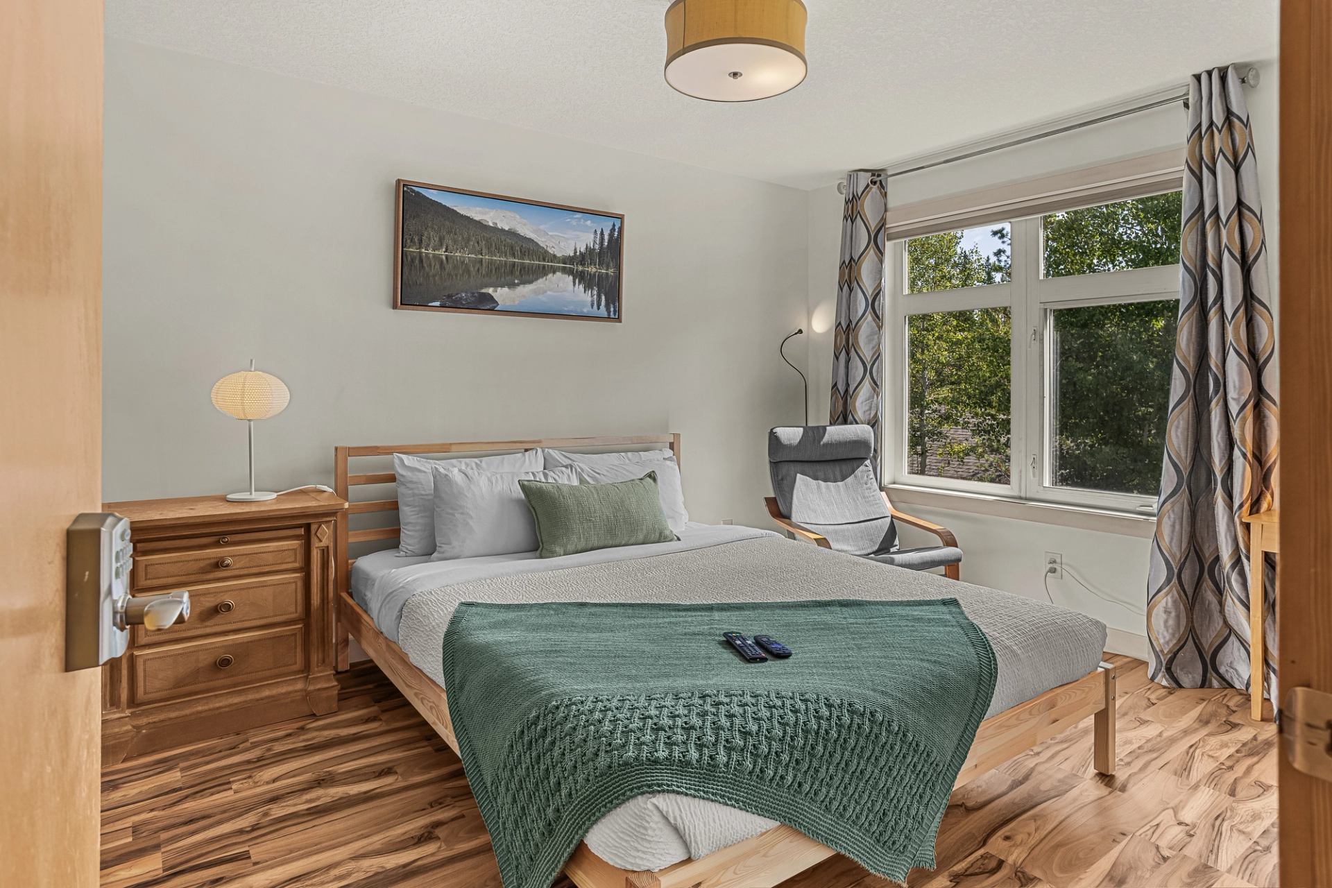 Bedroom with wooden bed, green throw, nightstand, armchair and large window.