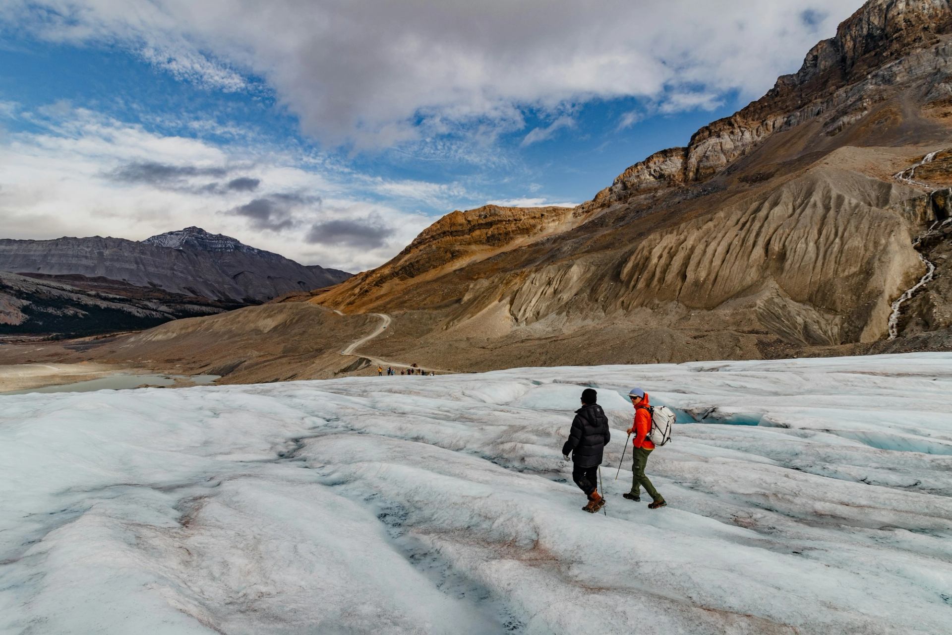 Hikers walk across a glacier with rocky mountains and cloudy blue sky behind.