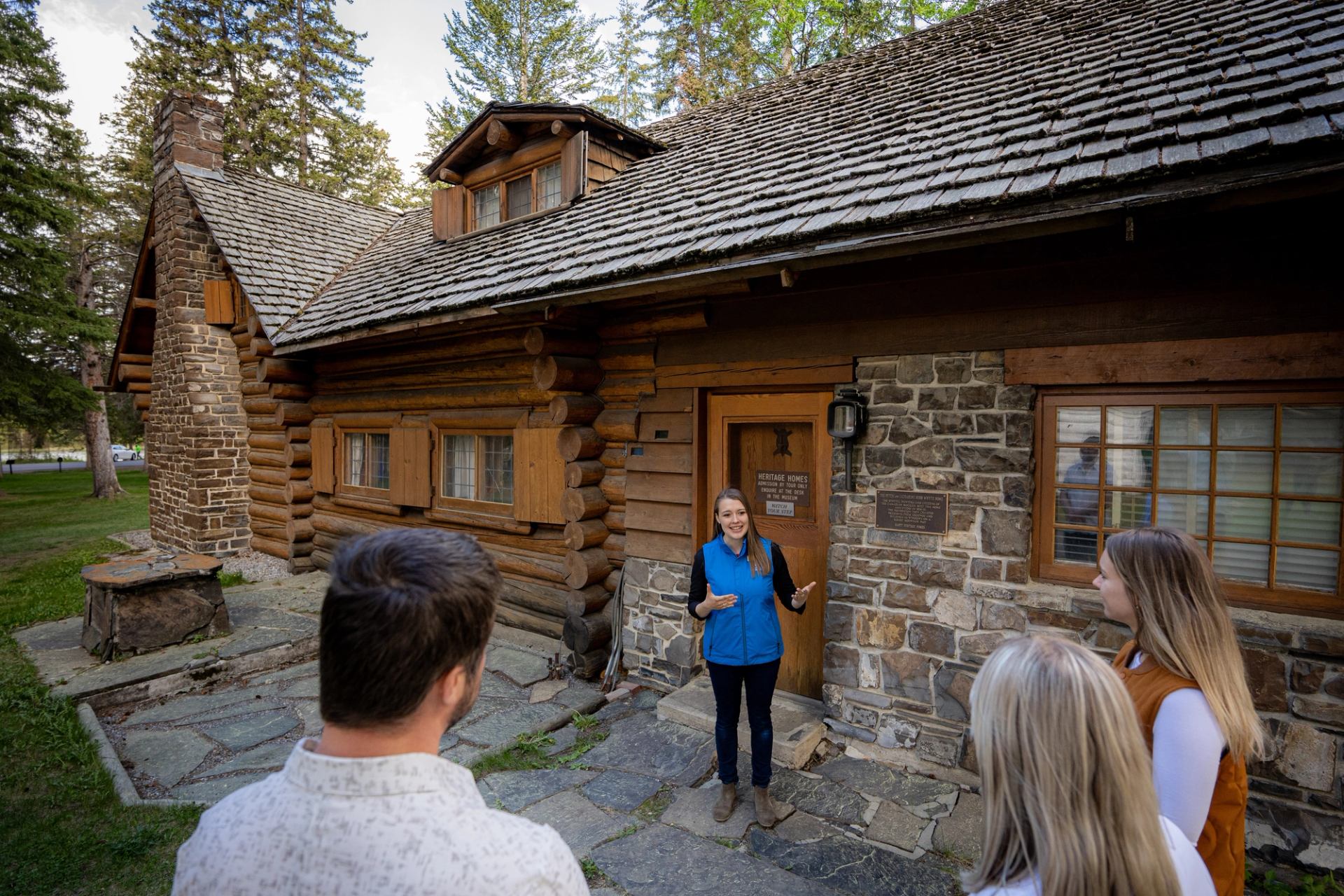 Guide speaks to visitors outside rustic log cabin with stone chimney and wooden door