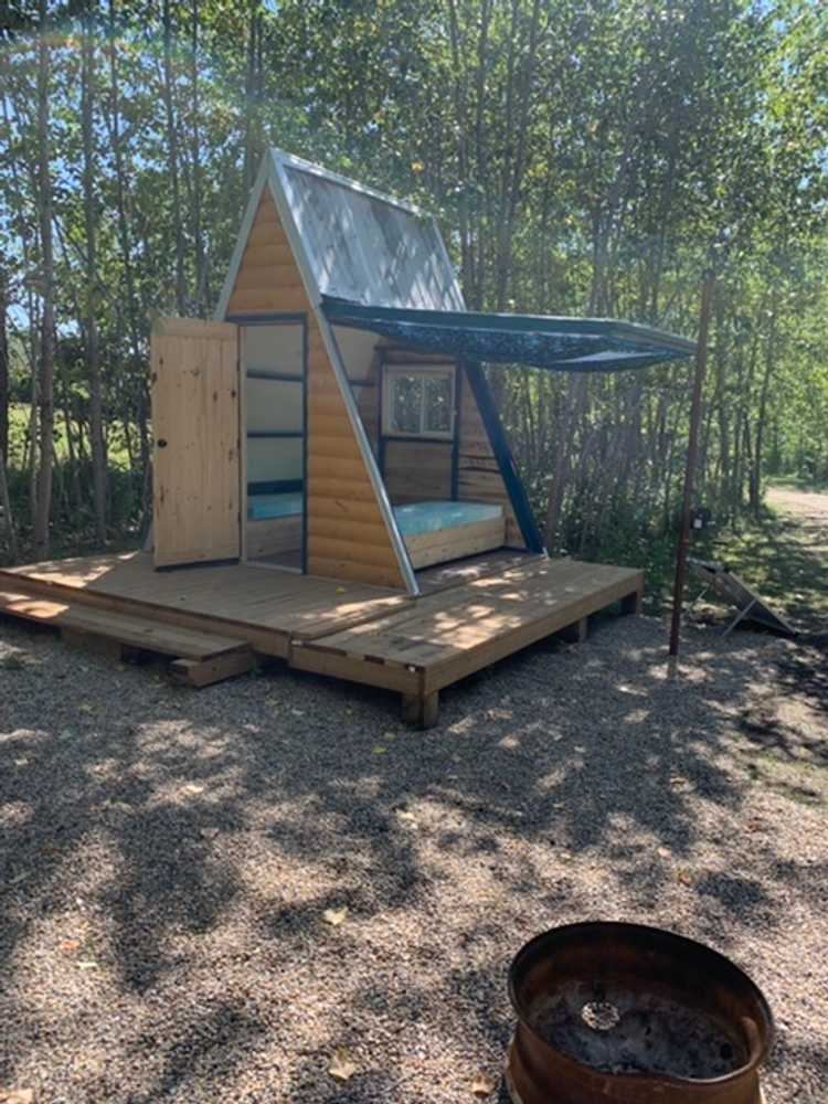 A small A-frame wooden cabin with an open door and two beds inside, on a raised deck with a fire pit, surrounded by trees.