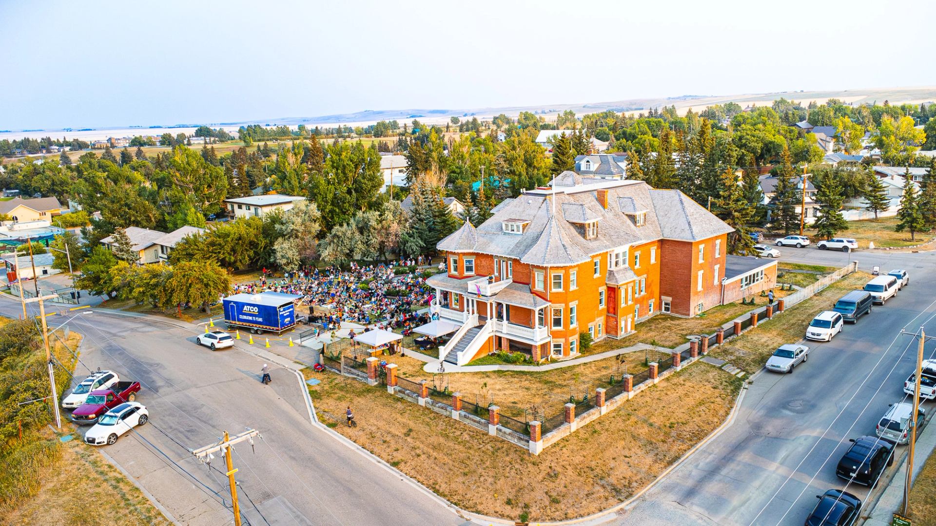 Large brick building on corner lot beside outdoor gathering and parked vehicles.