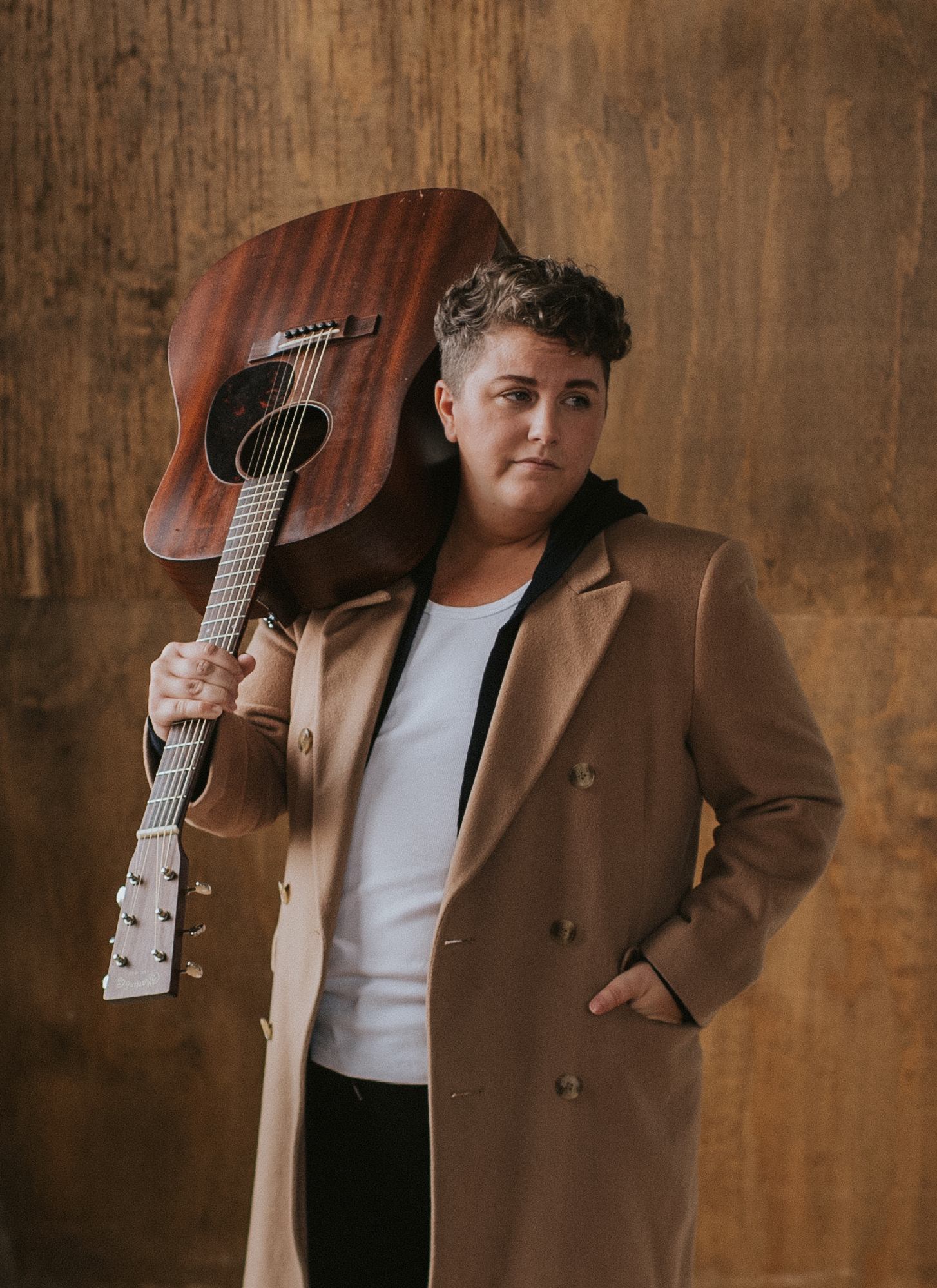 Person in tan coat holds acoustic guitar over shoulder against brown wall.
