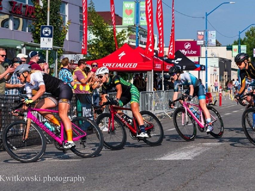 Cyclists race through a city street as spectators watch behind barriers and tents.