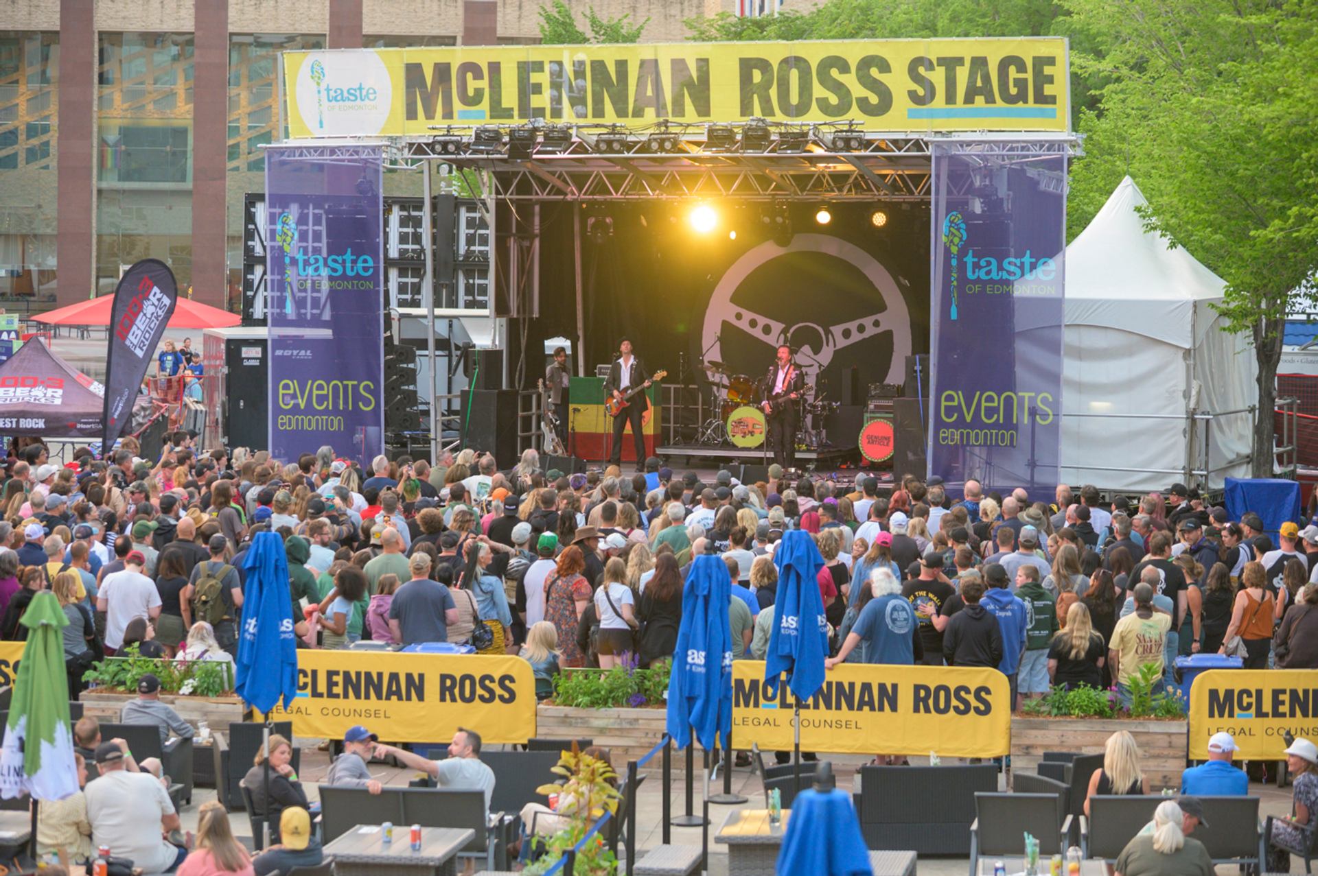 Band performing on a brightly lit outdoor stage before a large audience.