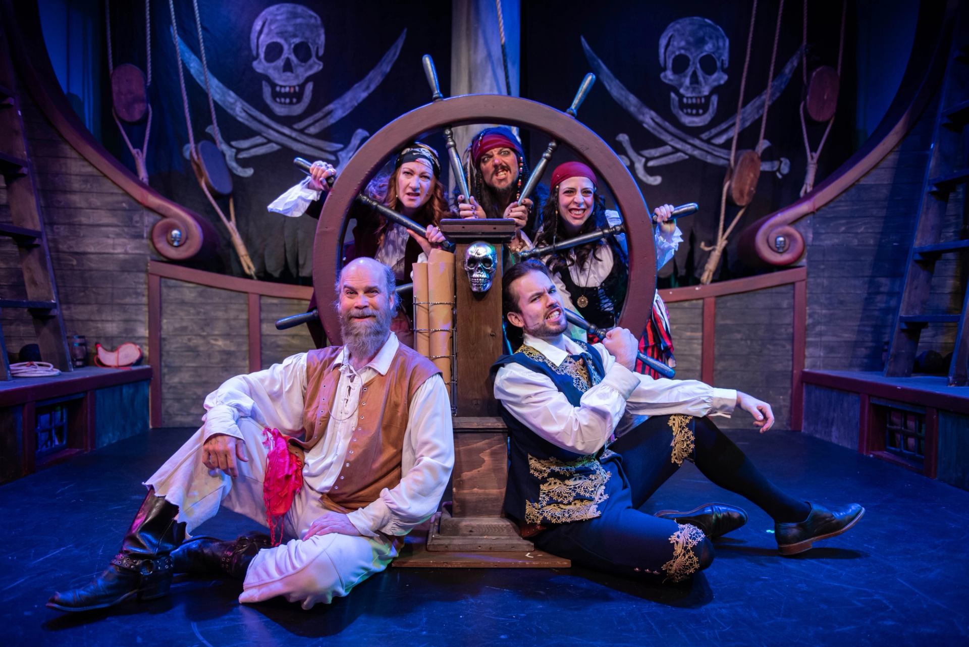 Actors in pirate costumes around a ship’s wheel on stage