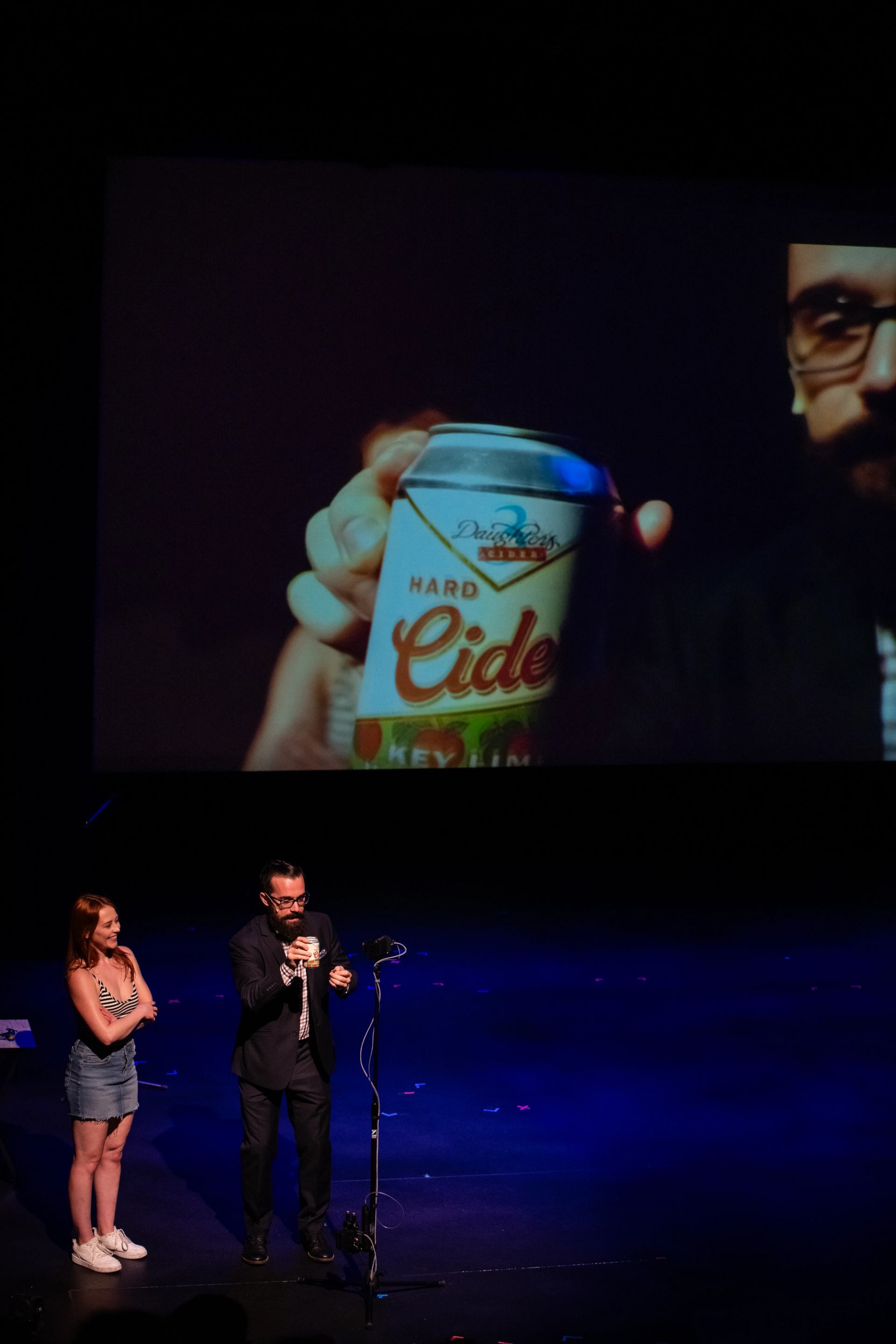 Stage performer holding a hard cider can beside volunteer, projected on large screen.