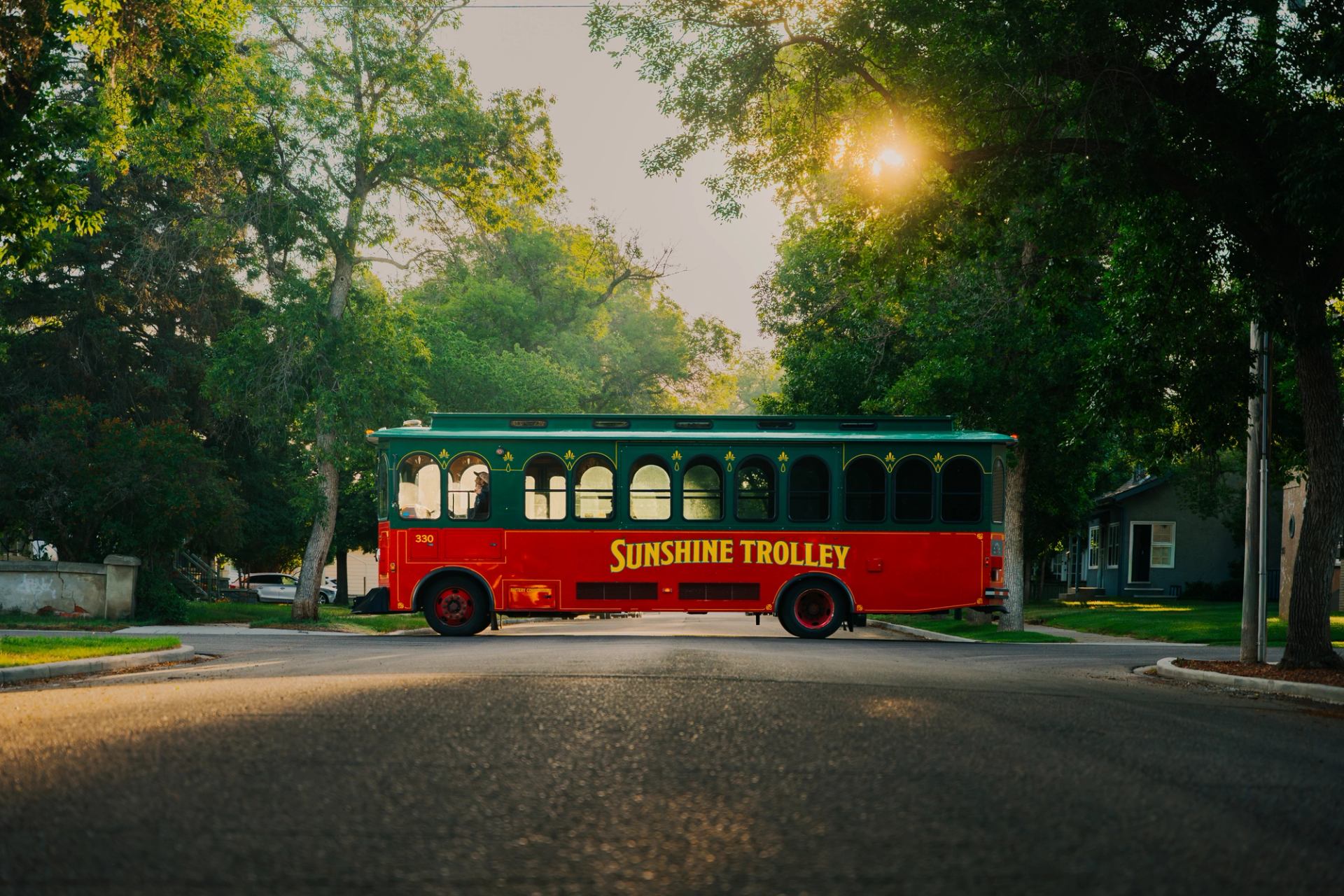 A red and green Sunshine Trolley traveling down a tree-lined street at sunset.