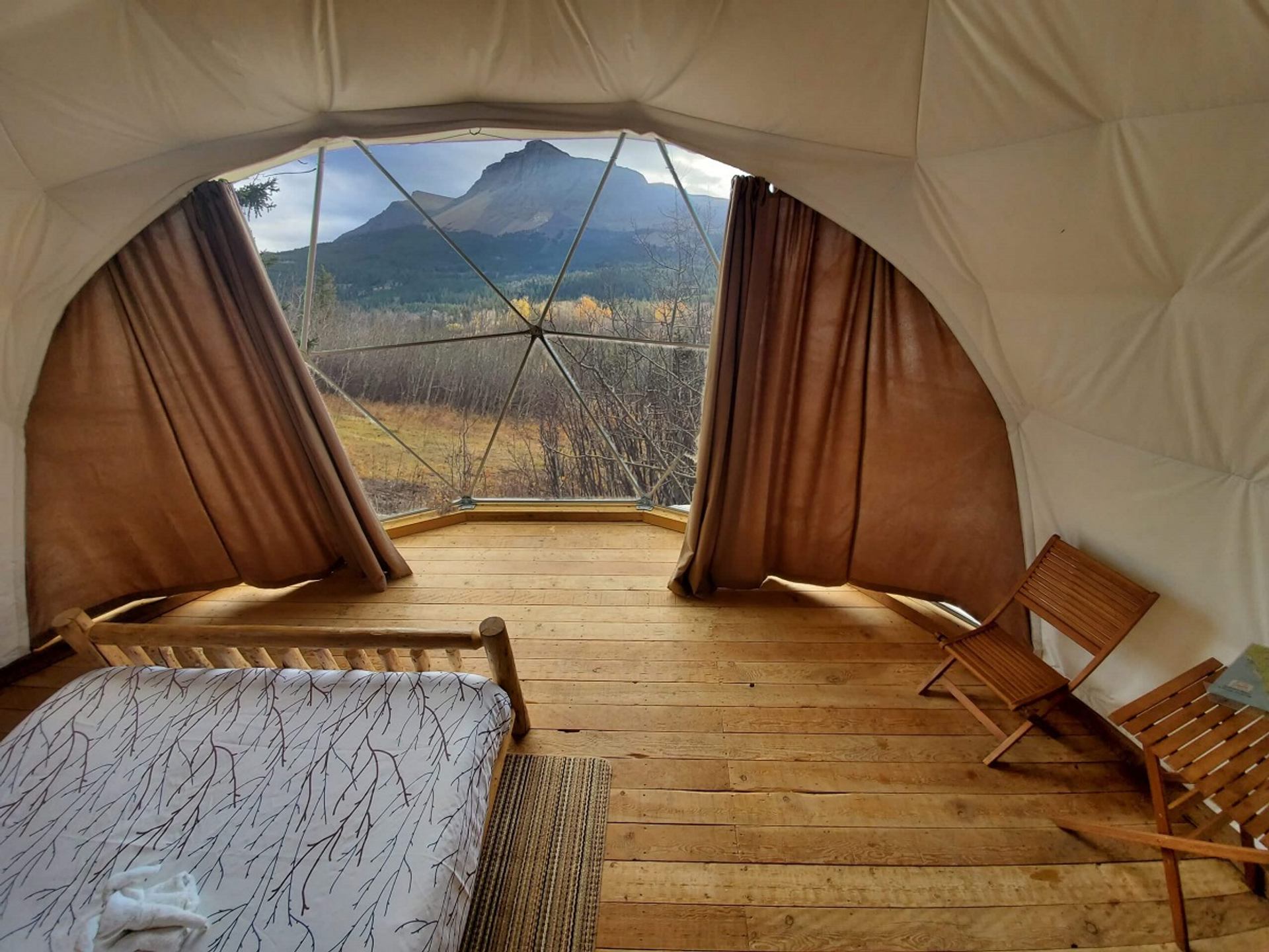 Glamping dome interior with wooden floor, bed, chairs, and panoramic mountain view.
