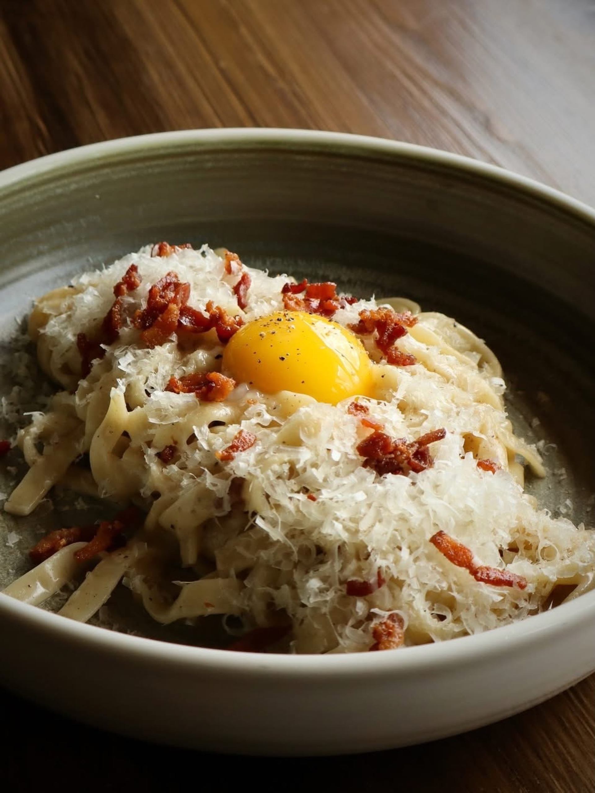 Bowl of pasta topped with raw egg yolk, grated cheese and bacon pieces.