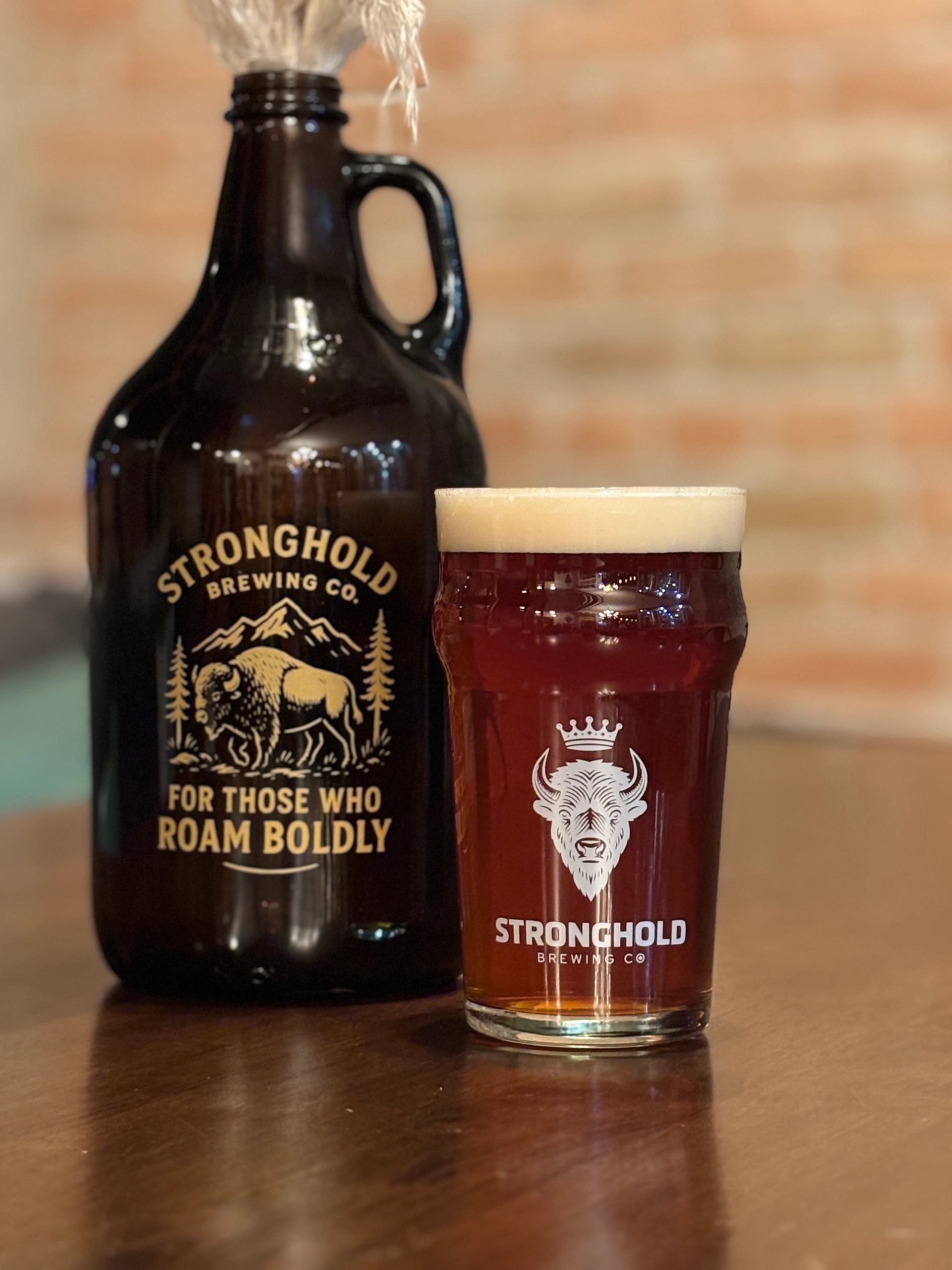 Glass of dark amber beer beside a Stronghold Brewing Co. growler on a wooden table.