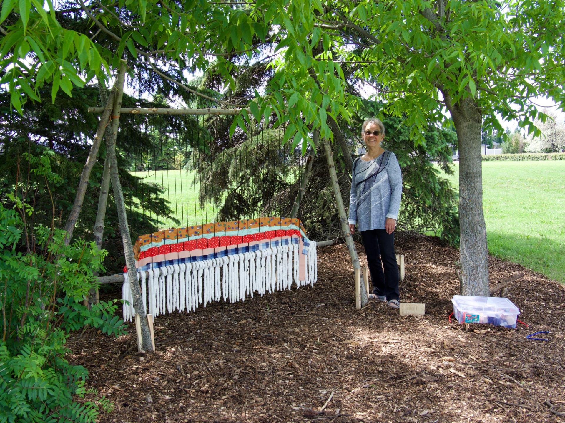 Person standing beside outdoor loom with colourful woven fabric under leafy trees.