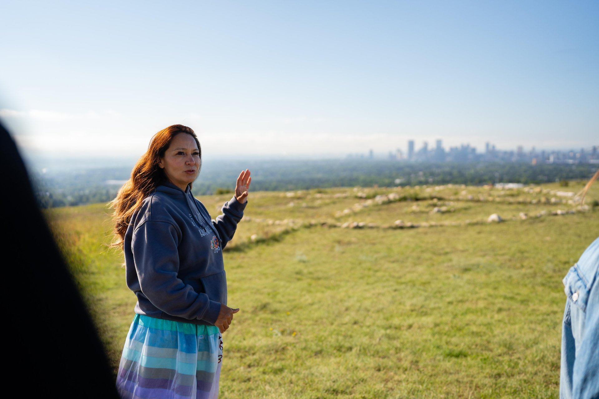 A person gestures across a grassy hill with a distant city skyline in view.