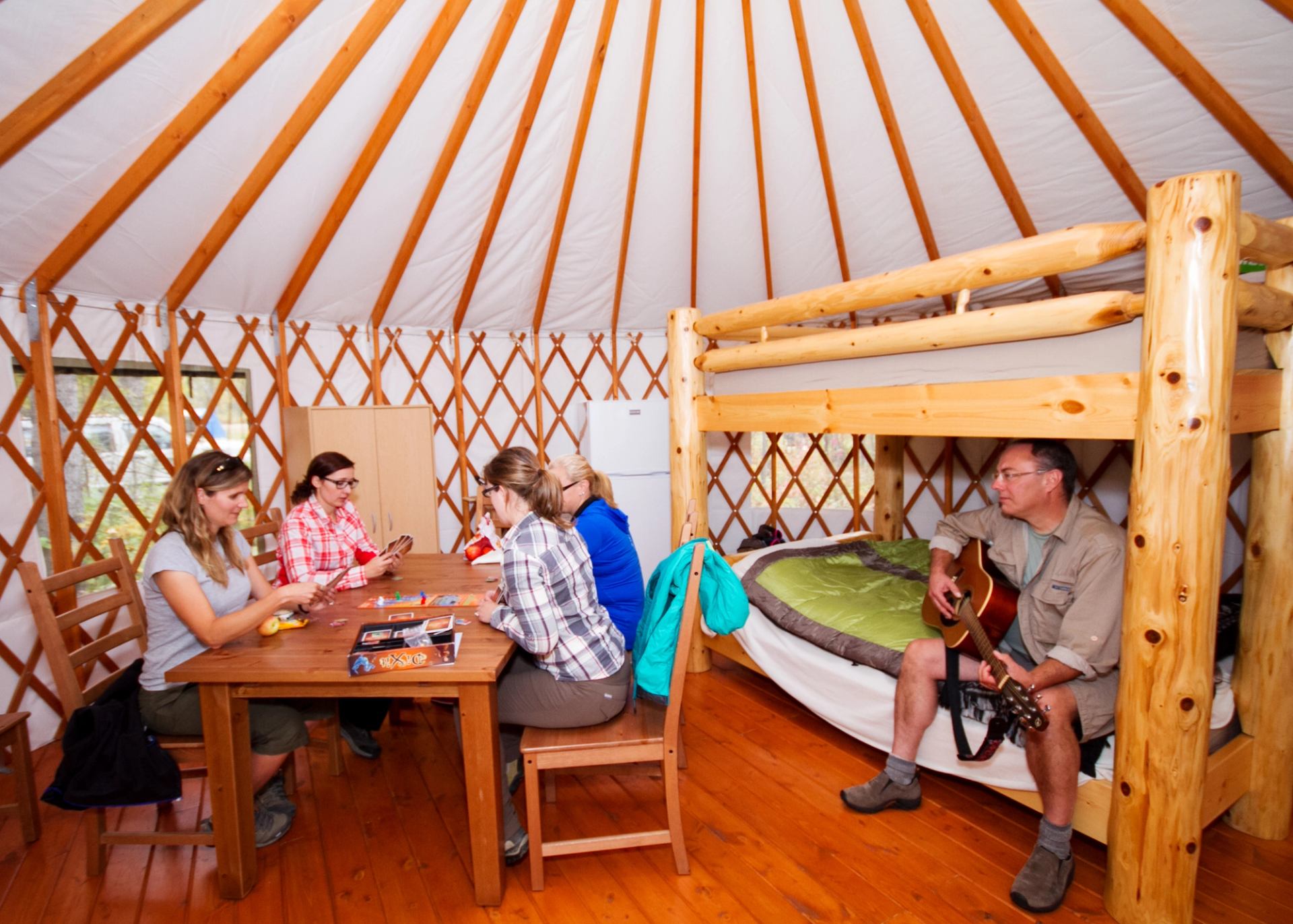 People sit at a table playing cards while another person plays guitar on bunk bed.