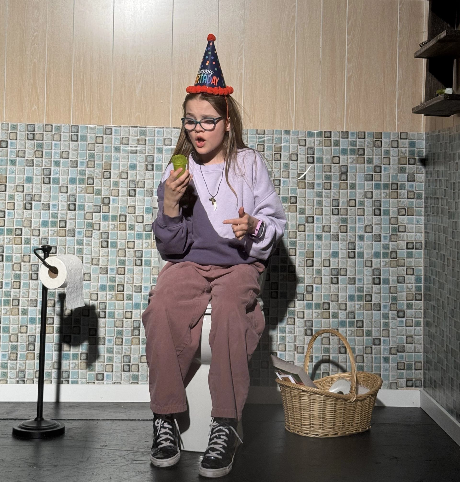 Person in party hat sitting on toilet, holding object in bathroom.