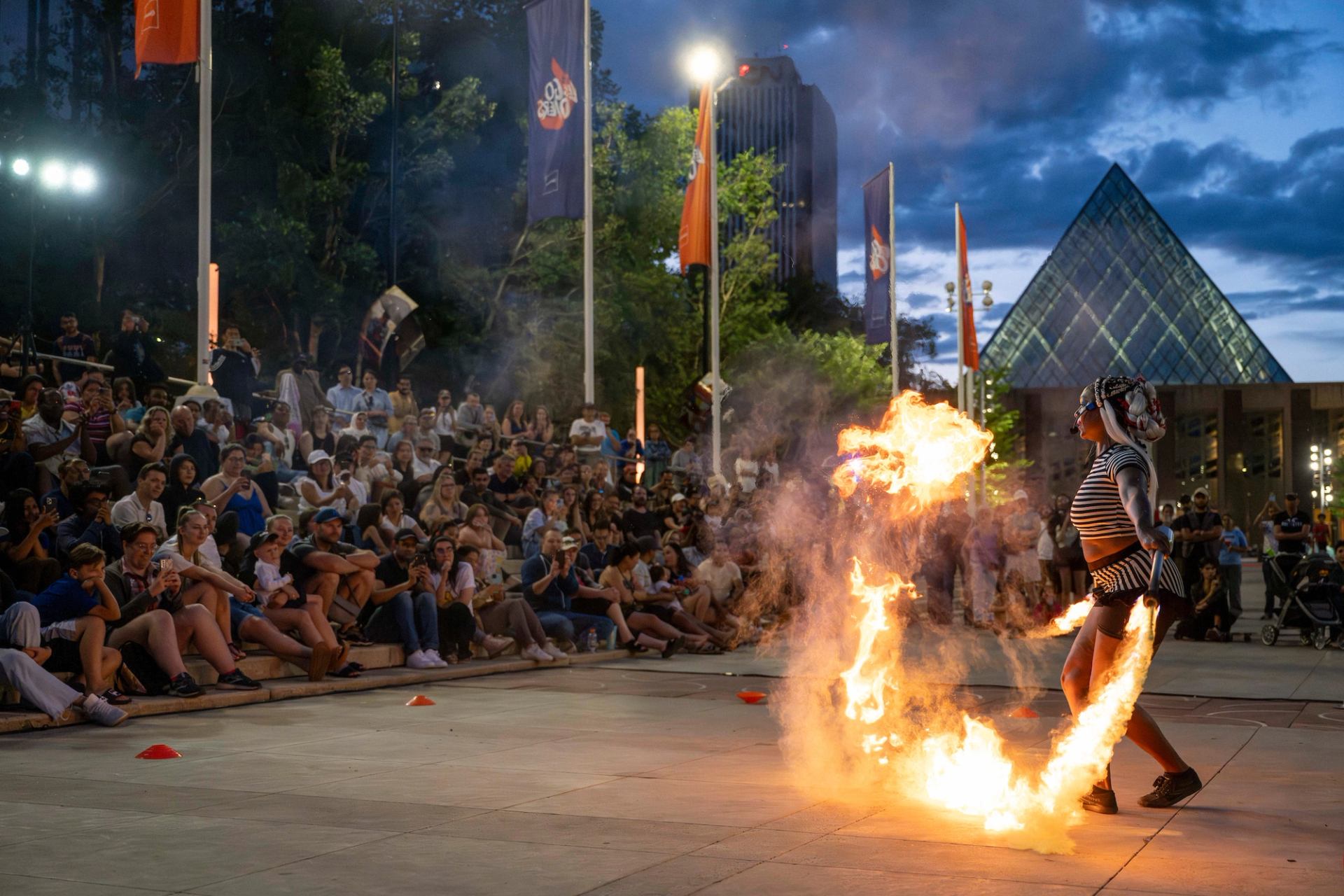 Fire performer swings flaming ropes before seated crowd in outdoor urban plaza.