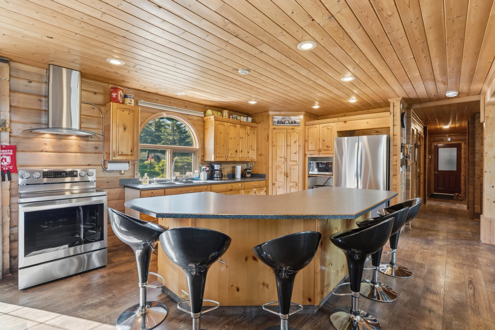 Wood-panelled kitchen with large island, black bar stools, stainless appliances and arched window.