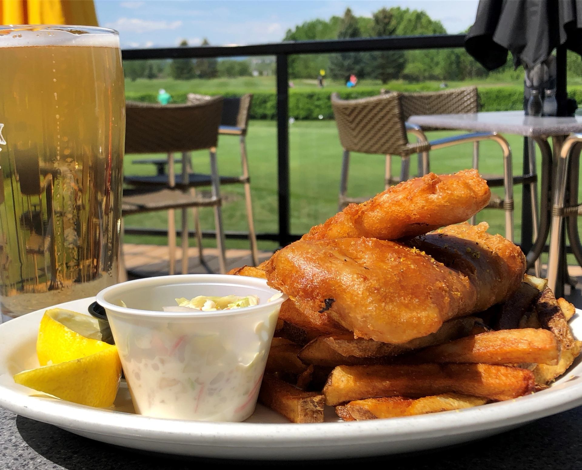 A plate of battered fish and fries served with coleslaw and a lemon wedge next to a full beer.
