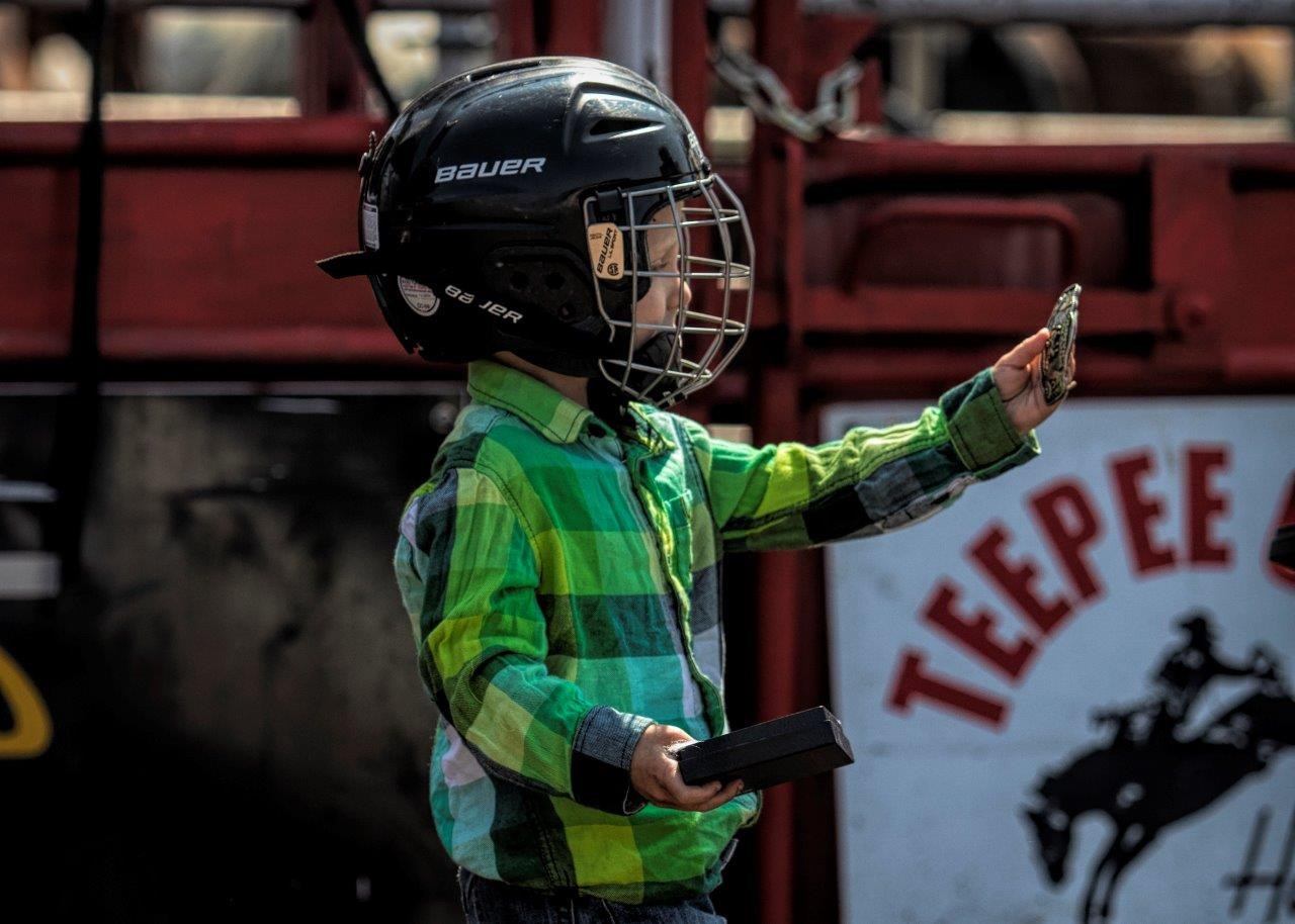 Child in plaid shirt wearing hockey helmet holds small object near rodeo sign.