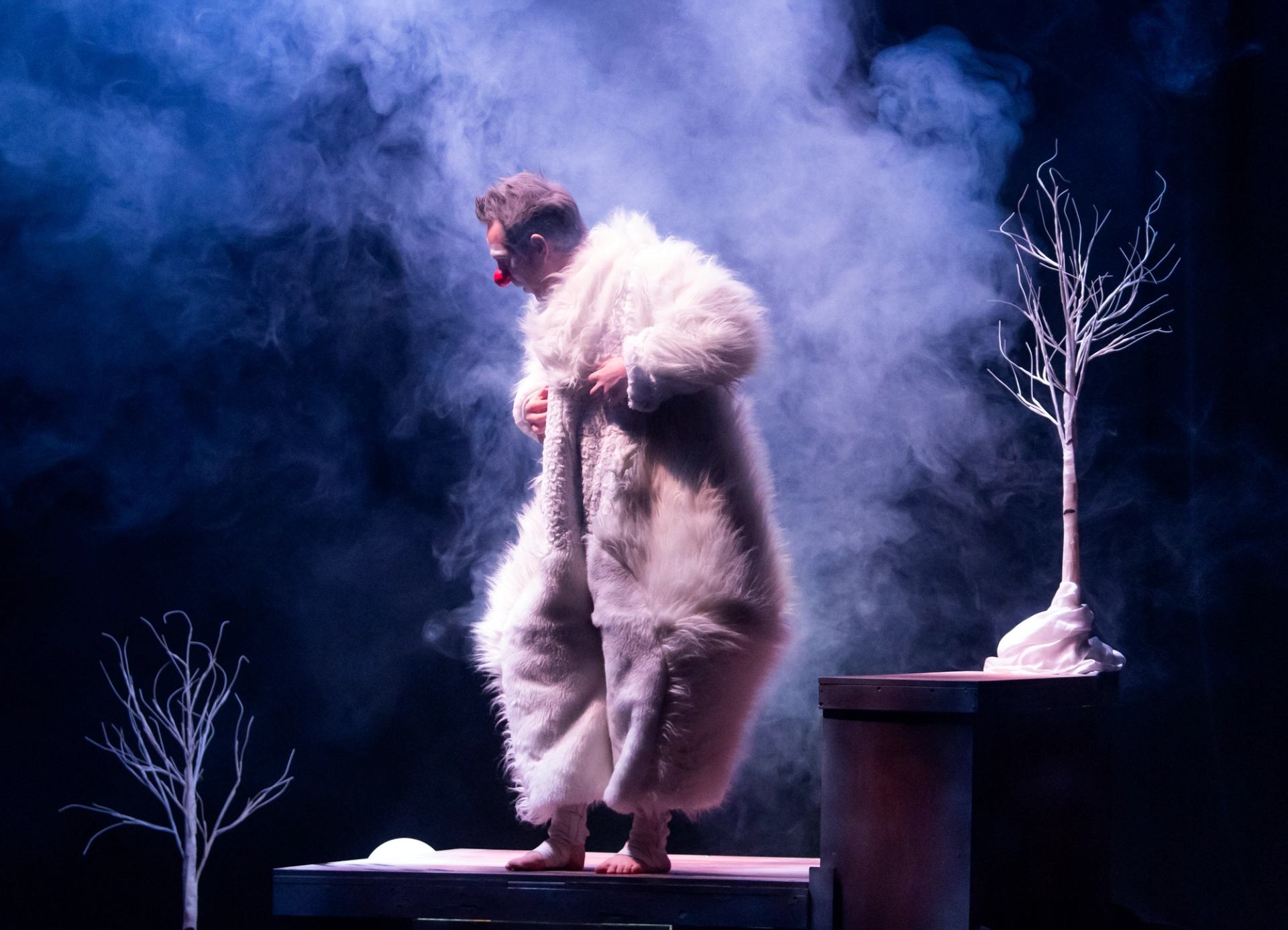 Stage performer in furry costume and red clown nose stands amid smoke and bare trees.