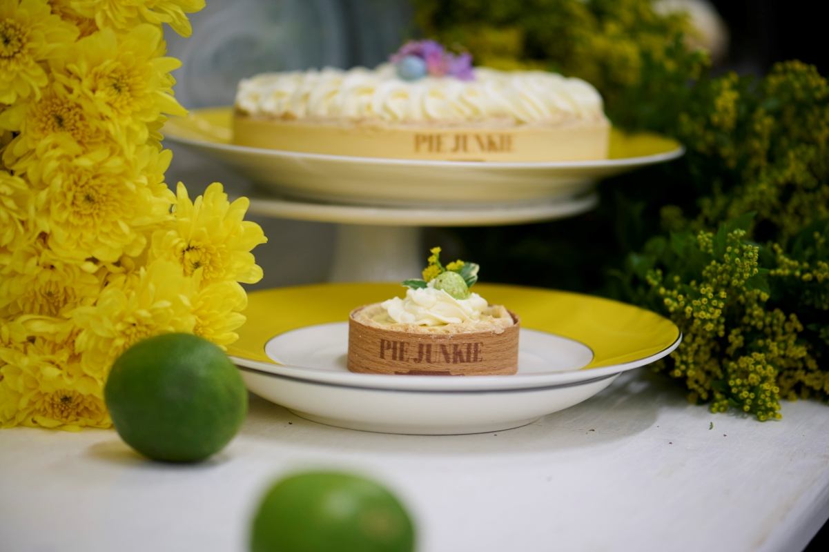 Two "Pie Junkie" tarts, one small and one large, with white cream toppings, displayed among bright yellow flowers, green foliage, and fresh limes.
