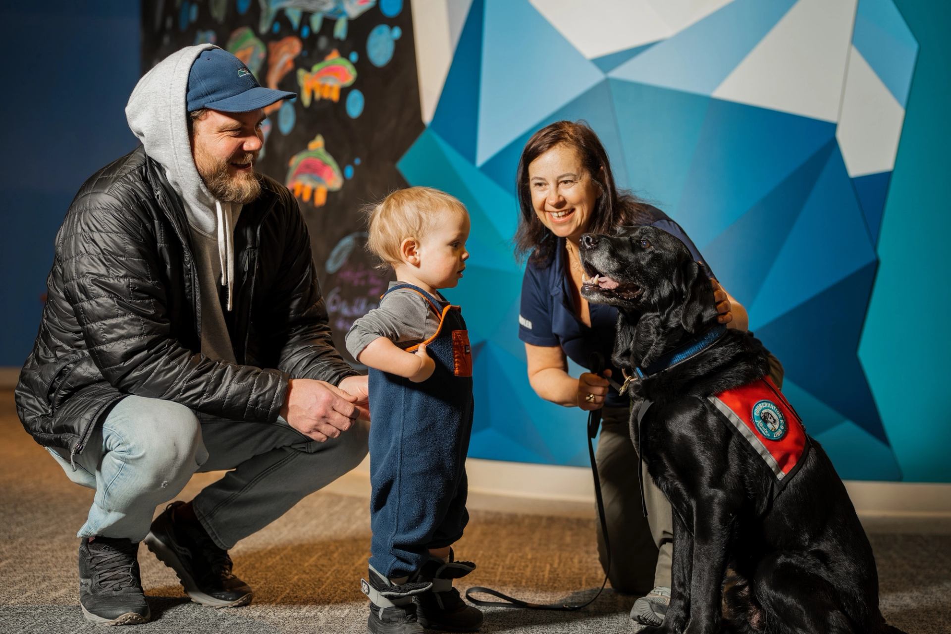 Child standing near a service dog with two adults at an indoor Fishtival event.