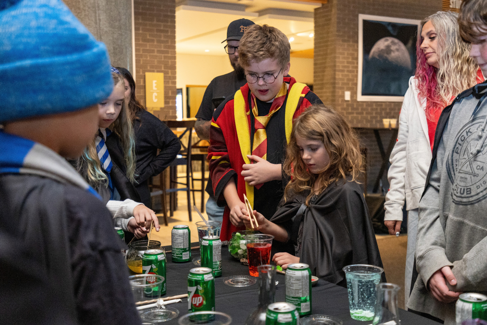 Costumed children mix colourful drinks at a table covered with cups and soda cans.