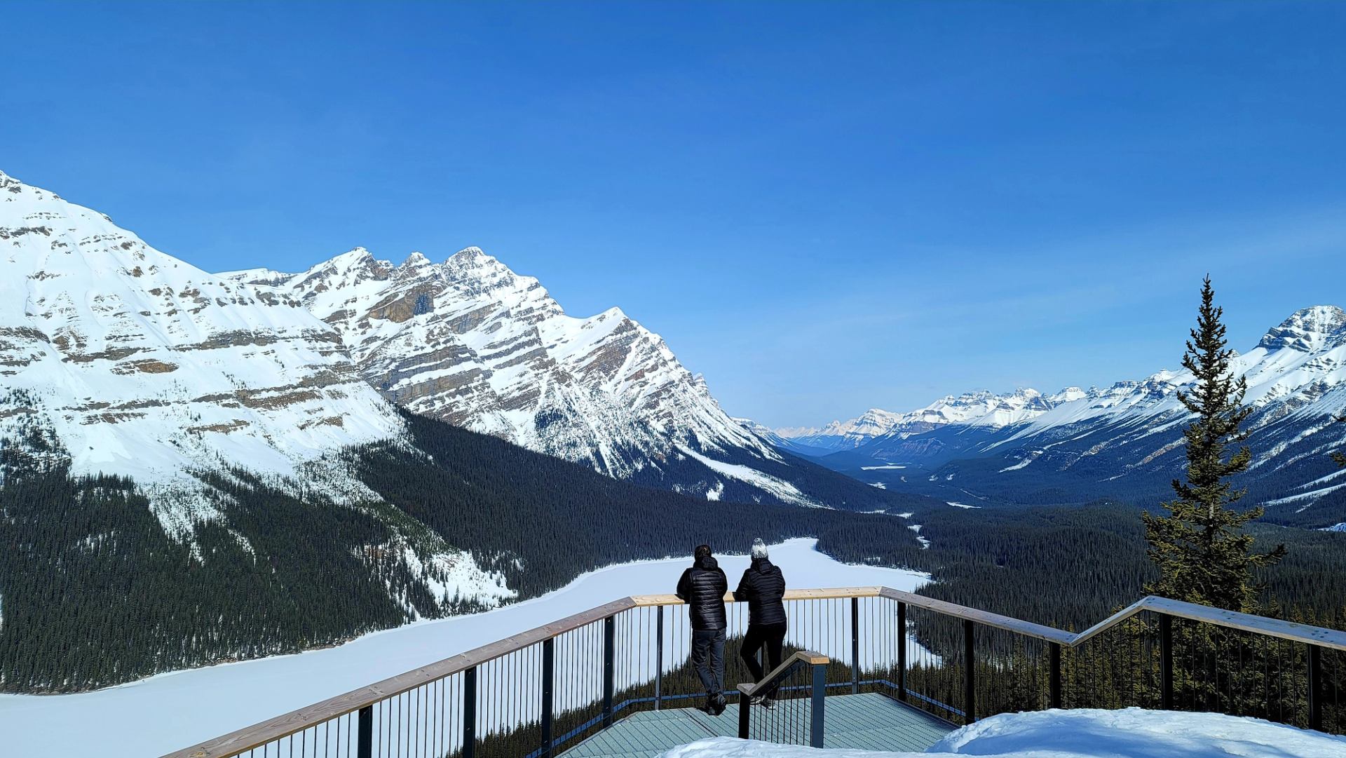 Two people standing on a snowy lookout deck overlooking a frozen valley and rugged mountains.